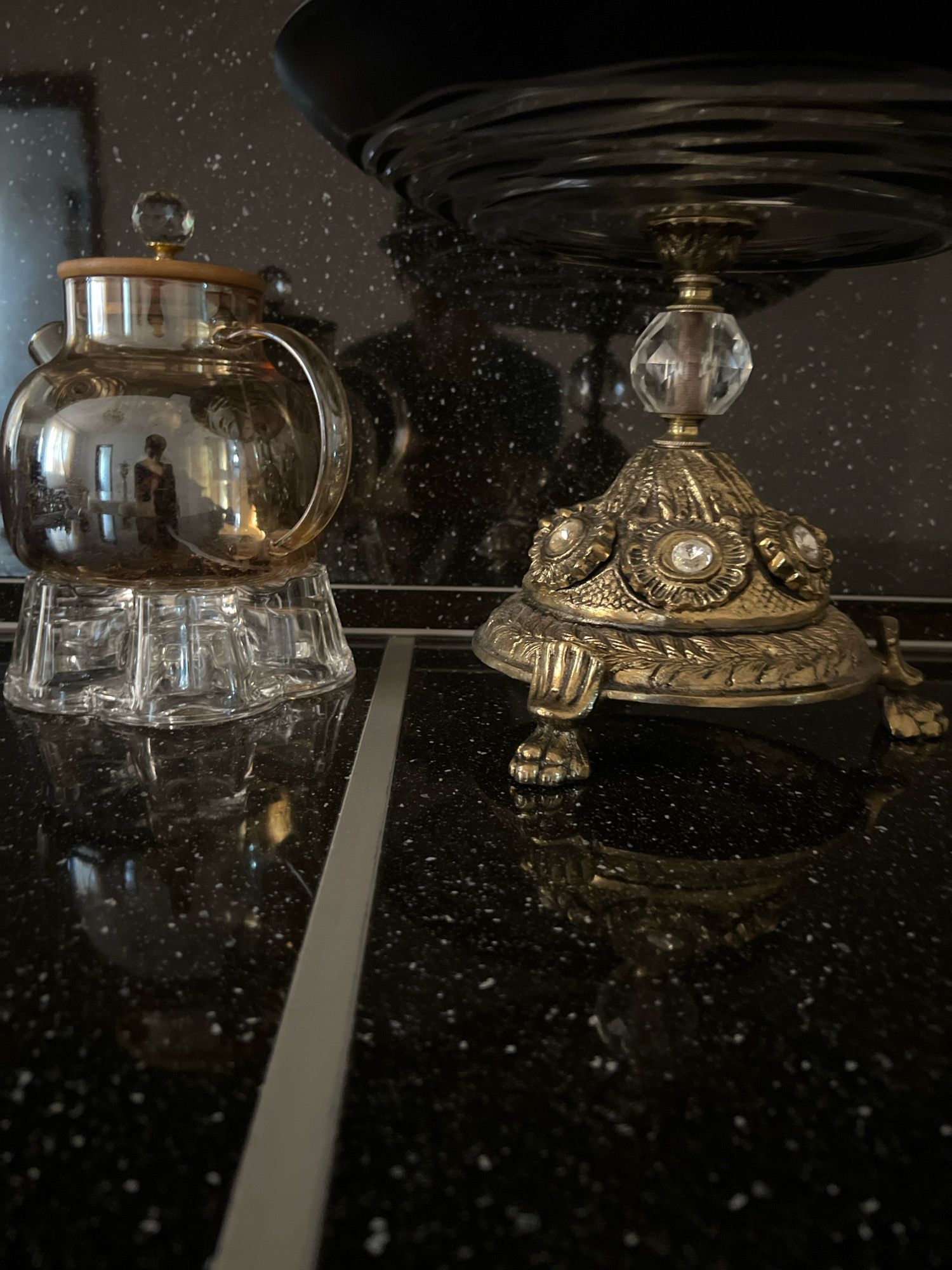 The image depicts two decorative objects placed on a black speckled countertop. On the left is a translucent, golden-colored glass teapot with a wooden lid topped by a clear glass knob. It rests on a clear glass base with a stepped, multi-faceted design. The teapot reflects a room with a person standing and windows. To the right is a brass decorative stand with intricate detailing, including what appear to be faux jewels and floral motifs. This stand has claw-like feet and supports a dark, possibly glass, bowl or platter at the top. The countertop has a reflective surface, showing reflections of the objects and the surrounding environment. A thin, light-colored strip runs vertically down the center of the image, dividing the two objects. The background is dark and also speckled, suggesting it is part of the countertop or a backsplash. The lighting is somewhat dim, casting reflections and shadows. There is no indication of the city of Novogagatli, Russia, in the image.