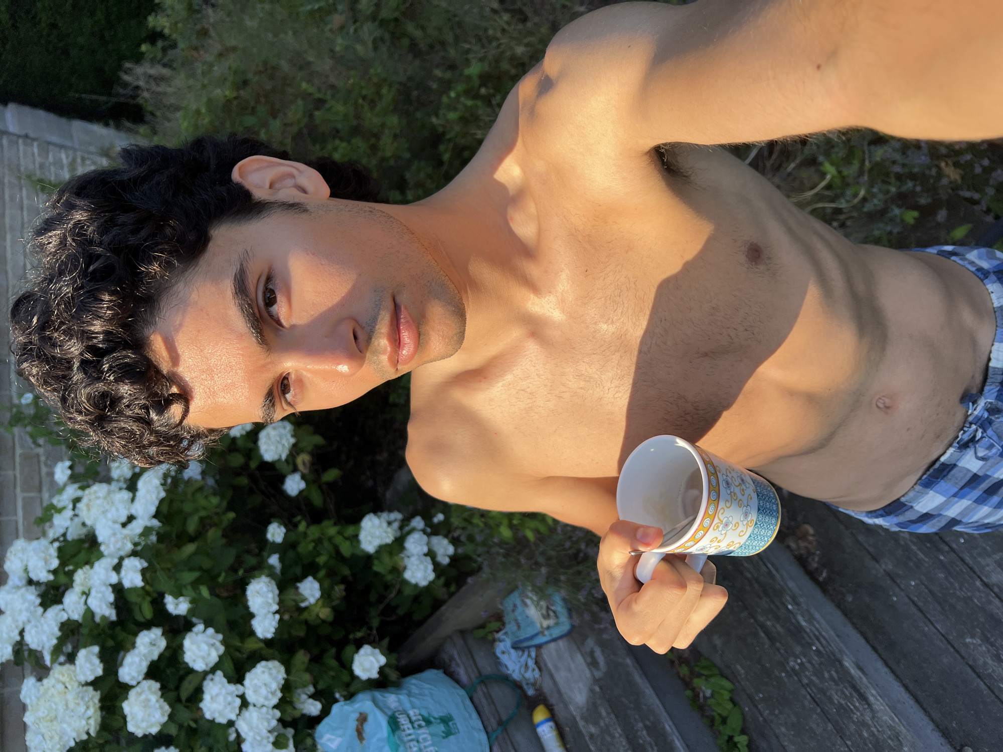 A young man, appearing to be in his early twenties, is captured in a selfie outdoors during daylight. He is shirtless and wearing blue and white plaid boxer shorts. He holds a white mug with a floral design in his right hand, and the mug contains a light-colored beverage. The man has dark, curly hair and his skin is illuminated by golden sunlight. He is standing on wooden planks, possibly a deck or stairs, with lush green foliage and white flowers in the background. A blue recycling bag is visible to the left of the man, along with a yellow bottle and a cloth. The setting suggests a residential backyard. The location is identified as Rotheux-Rimière, Belgium.
