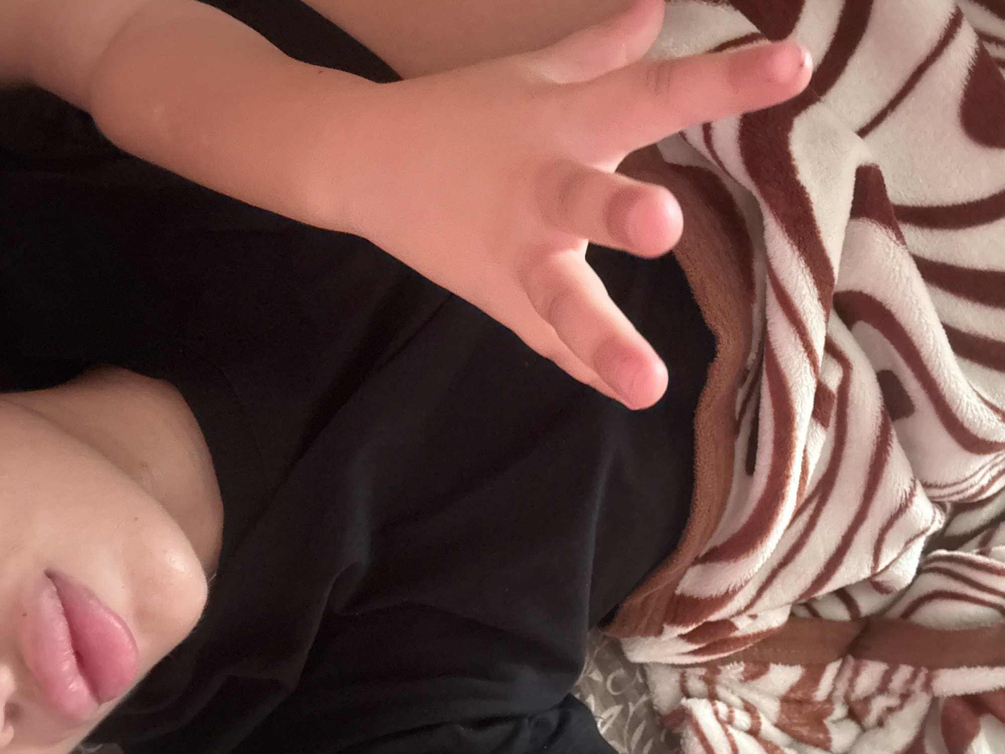 The image shows a close-up view of a person's face and hand, as well as a blanket with a brown and white striped pattern. The person's lips are pink and slightly parted, and their skin appears to have a warm tone. The hand is that of a child, with five fingers visible. The child's hand is reaching out towards the viewer. The background is a black garment, likely a t-shirt. The blanket is draped over what appears to be a bed or sofa. There is no indication of the location being Basarabeasca, Moldova.