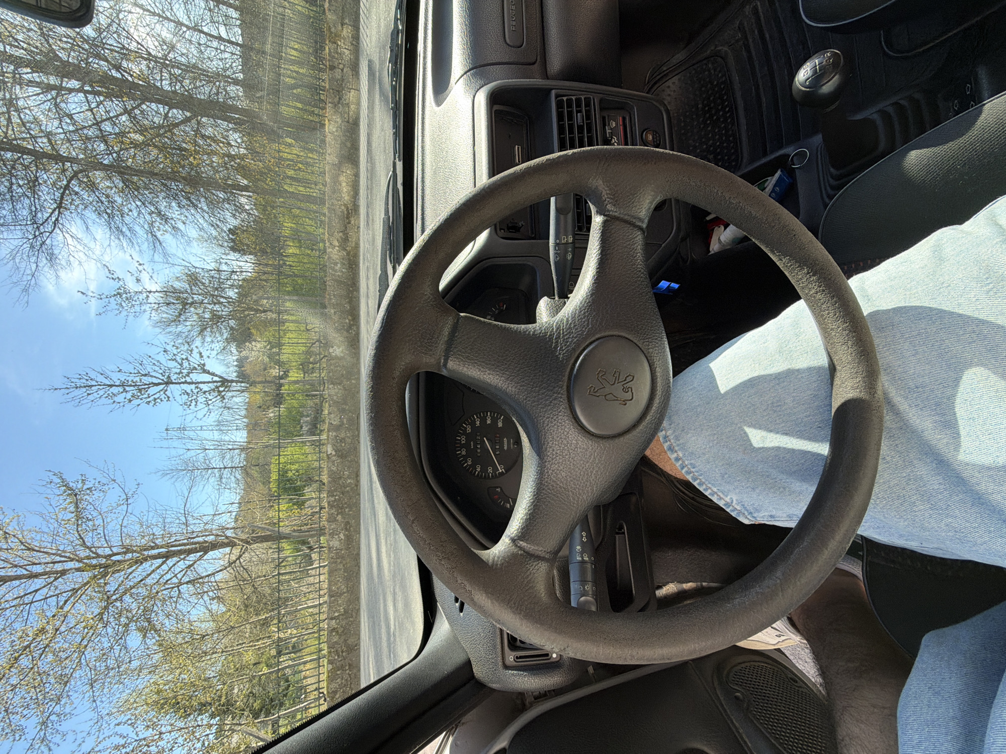 The image shows the interior of a Peugeot car, specifically the steering wheel, dashboard, and driver's side area. The steering wheel is black with the Peugeot logo at its center. The dashboard features an analog speedometer displaying 184128 kilometers. The driver, visible by their denim-clad legs and partially visible white sneaker, is in the driver's seat. The car's gear stick and black floor mats are visible. Through the windshield, a roadside scene is visible with trees lining the road, and a blue sky above. Based on the make of the car and the environment visible through the windshield, the photograph was taken in Vila Real, Portugal.