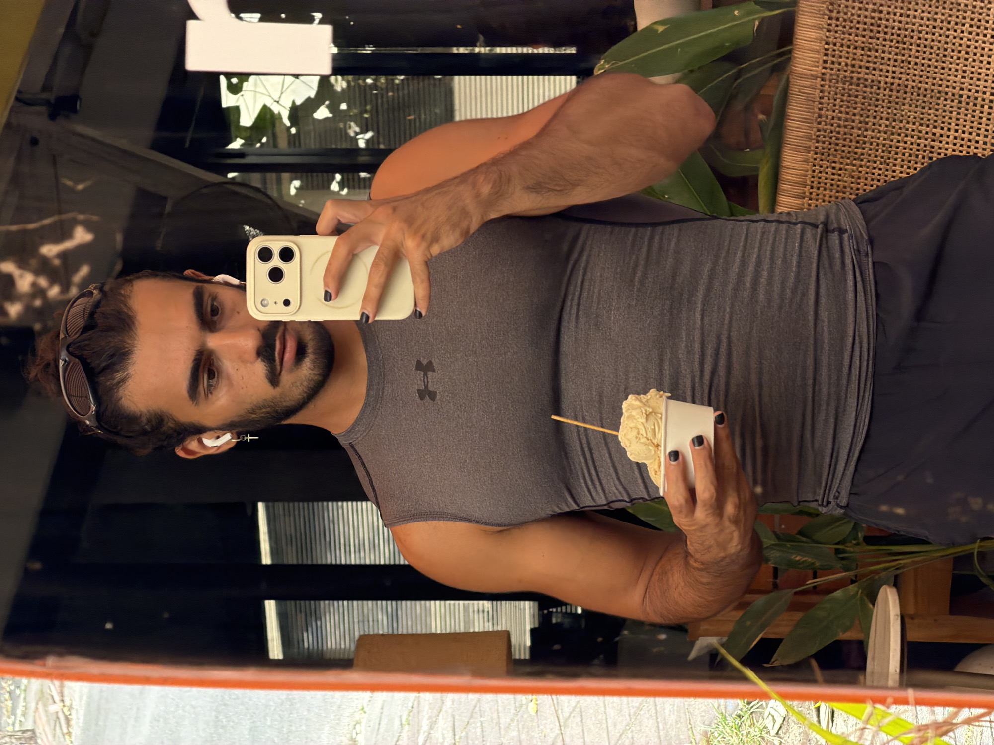 A man with dark hair tied in a bun and sunglasses perched on his head is standing in front of a mirror. He is wearing a grey sleeveless Under Armour shirt and black shorts. In his right hand, he holds a white iPhone 14 Pro, angled to capture his reflection. In his left hand, he holds a white cup with a light-colored ice cream, a wooden stick protruding from the top. The man has a beard and mustache, and is wearing white wireless earbuds, one with a cross-shaped charm hanging from it. His fingernails are painted black.
The reflection shows a background of blinds, a fan, and potted plants, suggesting an indoor setting possibly with an open-air component. The mirror itself has an orange frame on the left side of the image. The overall impression is a casual, everyday moment captured by the man himself. The scene appears to be in North Kuta, Indonesia, based on the provided information.