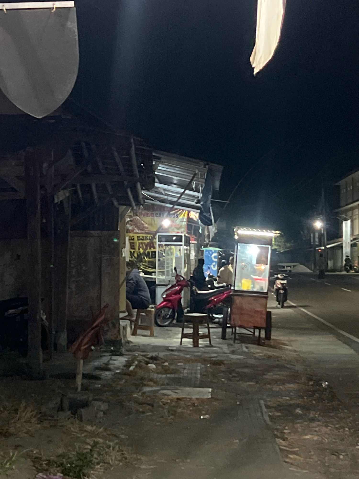 This is a night scene in Payaman, Indonesia, featuring a street-side food stall. A person sits on a stool next to a rustic wooden structure, possibly a shop or home. A red motorcycle is parked nearby. Further down the street, another food stall with a lit display case, possibly selling drinks or snacks, is visible, with several people gathered around it. A street lamp illuminates the area, casting long shadows. Motorbikes are also seen parked on the side of the road. The overall environment appears to be a typical urban setting at night, with evidence of local commerce and daily life.