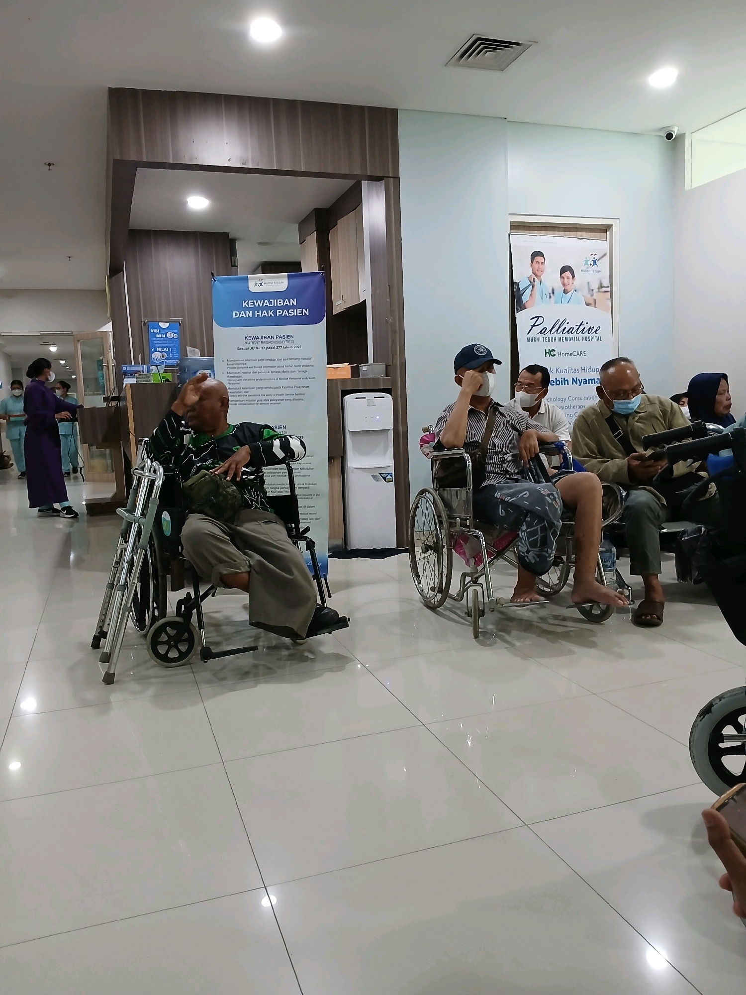 The image shows an indoor scene, likely a hospital waiting area, in the City of Medan, Indonesia. Several individuals are present, some in wheelchairs, suggesting they are patients.
In the foreground, a man is seated in a wheelchair, facing left, with his hand raised to his face. He is wearing a green and black patterned shirt and light-colored pants. To his right, another man in a wheelchair, wearing a striped shirt, a dark cap, and a mask, is looking down and appears to be touching his face. Further to the right, a man in a tan jacket and mask is looking at his phone. Behind him, a woman wearing a headscarf and mask is also seated.
In the background, three individuals in blue scrubs and masks are walking past a doorway on the left. A large blue and white banner with text in Indonesian is visible on the right, advertising "KEWAJIBAN DAN HAK PASIEN" (Patient Responsibilities and Rights) and mentioning "PALLIATIVE" services from "MURNI TEGUH MEMORIAL HOSPITAL". A white water cooler is also visible to the right of the banner. The floor is tiled with a glossy finish, reflecting the overhead lighting. The environment appears clean and well-lit, typical of a healthcare facility.
