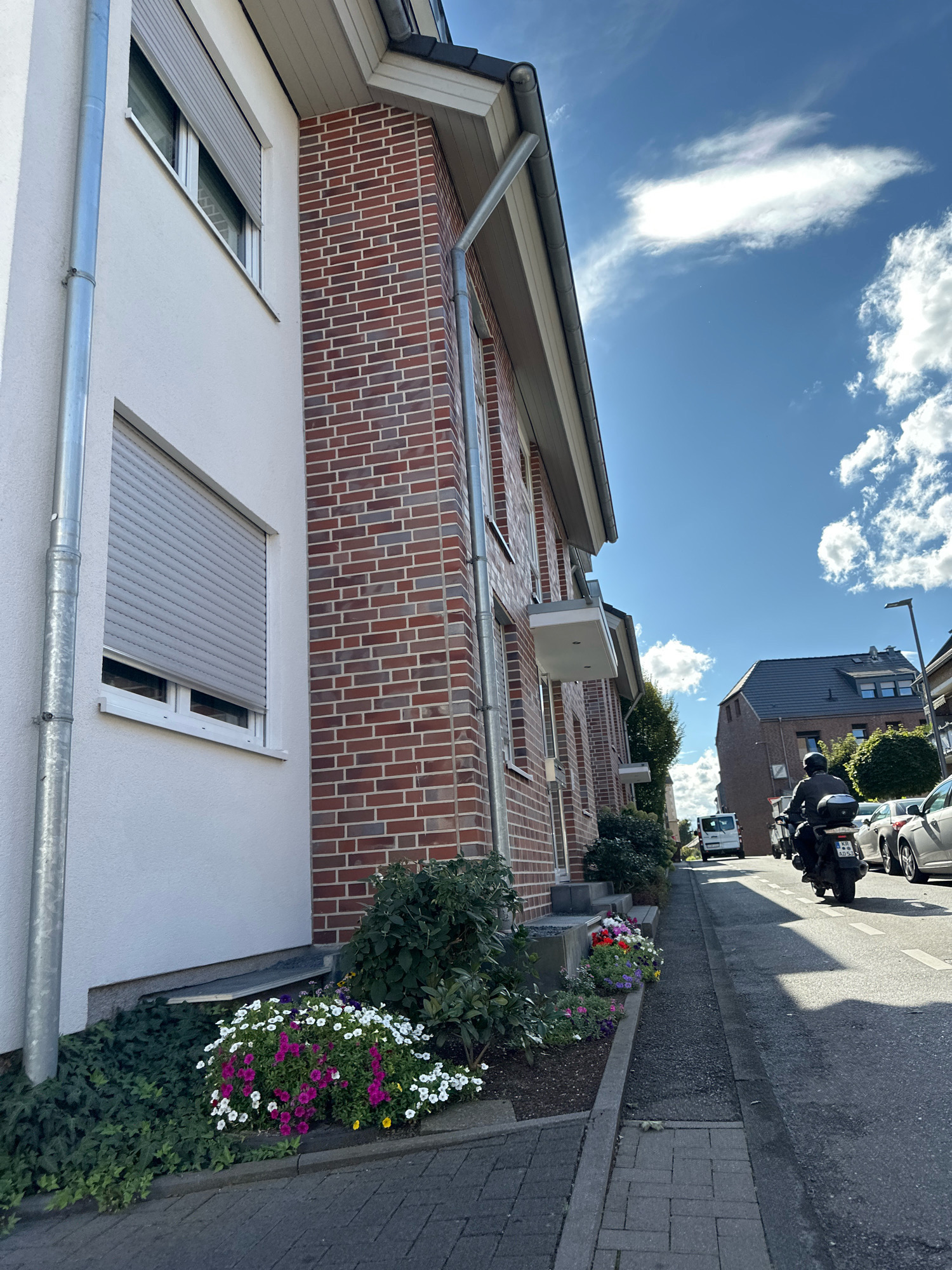 The image depicts a street scene in Krefeld, Germany, on a sunny day with scattered clouds. In the foreground, a white plastered building with red brick accents and a gray metal downspout is visible on the left. A window with a gray roller shutter is prominently featured. In front of the building, a flower bed with a variety of colorful flowers, including pink, white, and yellow, is planted along the sidewalk.
The street runs to the right, with a paved sidewalk and road. A black motorcycle with a rider wearing a helmet and a black backpack is moving away from the camera down the street. Several cars are parked along the right side of the street. Further down the street, a white van is visible, along with other buildings. Trees and greenery are present along the street and between the houses, suggesting a residential area. The overall impression is of a well-maintained urban environment.