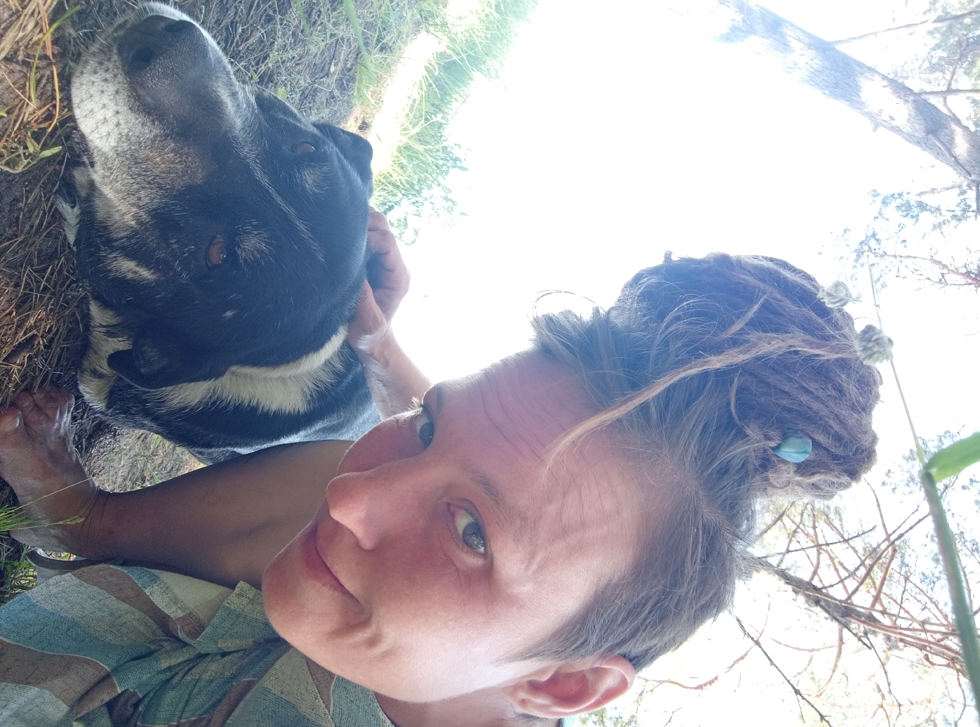 A selfie is taken from a low angle, showing a person with dreadlocks adorned with a blue bead and a small fluffy ornament. The person is looking towards the camera with a slight smile. To the left, a black and white dog is lying down, with the person's hand resting on its head. The background consists of dry grass, dirt, and the trunk and branches of a tree, with a bright sky overhead, suggesting an outdoor setting. The image was taken in Podborov'e, Russia.