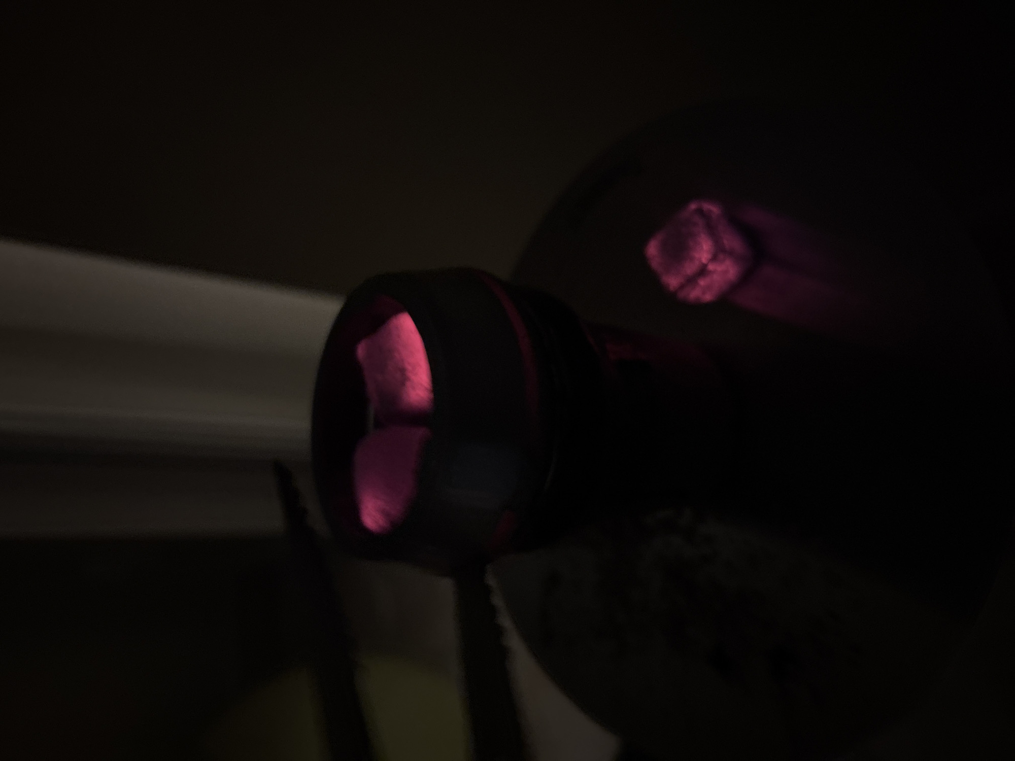 The image shows a close-up of a hookah setup in a dimly lit environment. Several glowing red coals are visible within the hookah bowl, indicating they are lit and hot. One detached coal with a similar glow sits to the right of the bowl on a dark surface, reflecting pink light. The background is dark and blurred, with a vertical element that could be a curtain or a wall. There are no people visible in the image, and no specific location in Krasnodar, Russia can be identified from the visual information provided. The scene appears to be indoors, possibly in a home or a lounge.