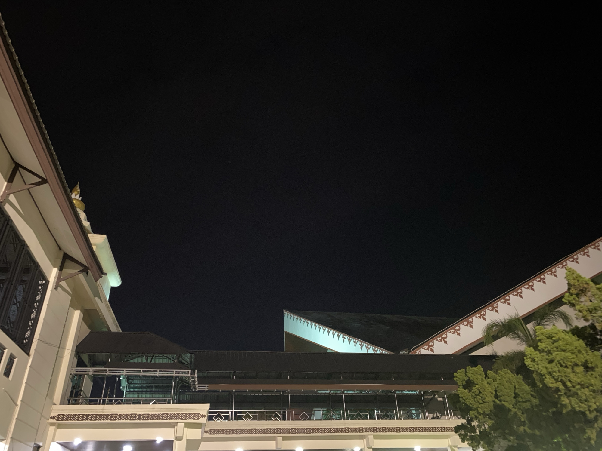 The image captures a nighttime view of modern architecture illuminated against a dark sky. The scene is dominated by the geometric shapes of buildings, with a focus on roofs and upper stories. A multi-level structure with dark roofing and balconies is visible in the mid-ground. To the left, a lighter-colored building with intricate window detailing and a decorative roofline is partially visible. The right side of the image features a building with a white facade adorned with a red and white patterned trim, sloping upwards. Lush green trees are present in the foreground on the right, partially obscuring the building. The overall impression is one of urban night scenery with a blend of traditional and contemporary architectural elements.
