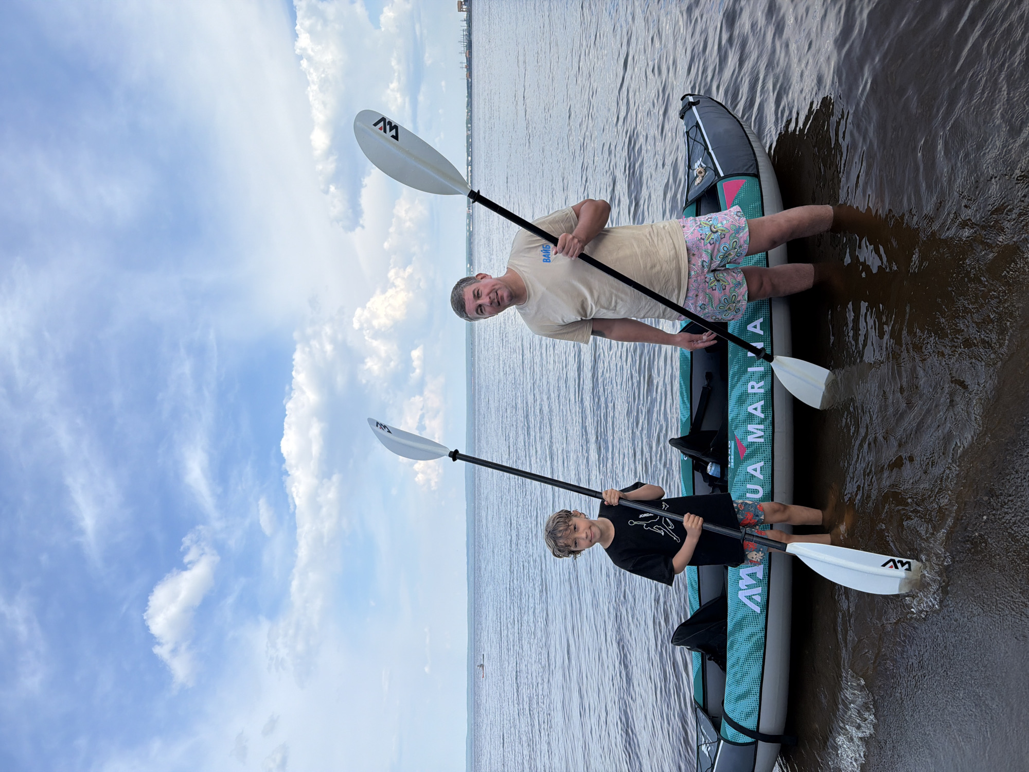 An outdoor, eye-level, full shot shows a man and a young boy standing in shallow water near a grey and teal inflatable kayak. The man, on the right, is wearing a light tan t-shirt with blue lettering and pink patterned shorts. He holds a white kayak paddle with a black shaft. The boy, on the left, wears a black t-shirt with a white graphic and patterned shorts. He also holds a white kayak paddle with a black shaft. The kayak has "AQUA MARINA" written on its side. The water is calm with small waves lapping at the shore. The sky is blue with scattered white clouds. In the distance, there is a body of water with a faint shoreline and what appears to be industrial structures. The location is the bank of a river in Noviy, Russia.