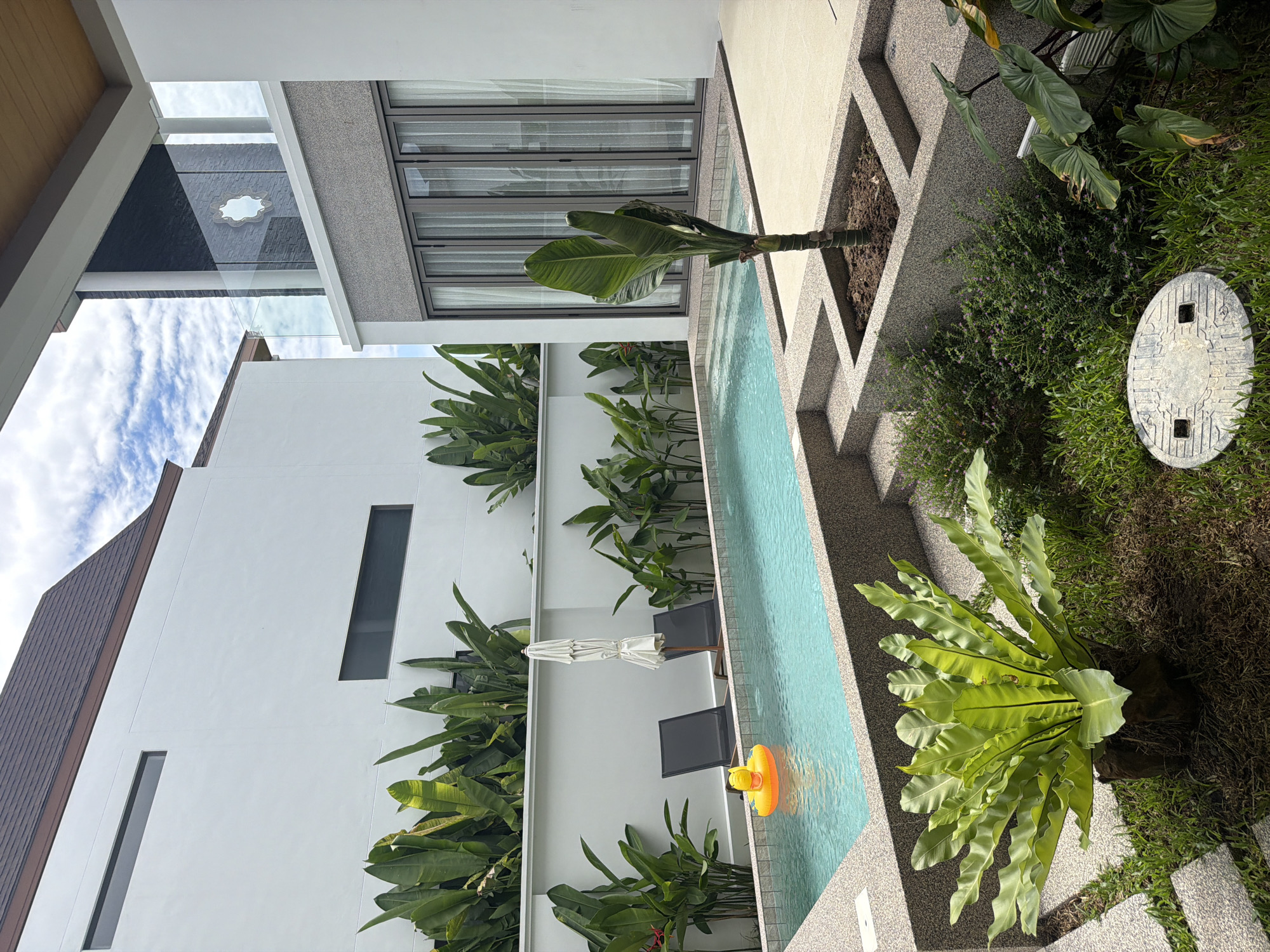 The image depicts a serene outdoor swimming pool area at a modern villa in Thep Krasatti, Thailand. The pool, with its turquoise water, is surrounded by a textured concrete deck and planters. A bright yellow duck-shaped inflatable toy floats near the shallow end, where two dark lounge chairs and a closed white umbrella are positioned. Lush green tropical plants, including large-leafed bird's nest ferns and banana-like foliage, frame the pool area and the adjacent stark white walls of the villa. One wall features a horizontal slit window. The villa itself has a dark tiled roof and a modern facade with large glass doors. The sky above is a mix of blue and white clouds. The scene is tranquil and suggests a private, luxurious residence.