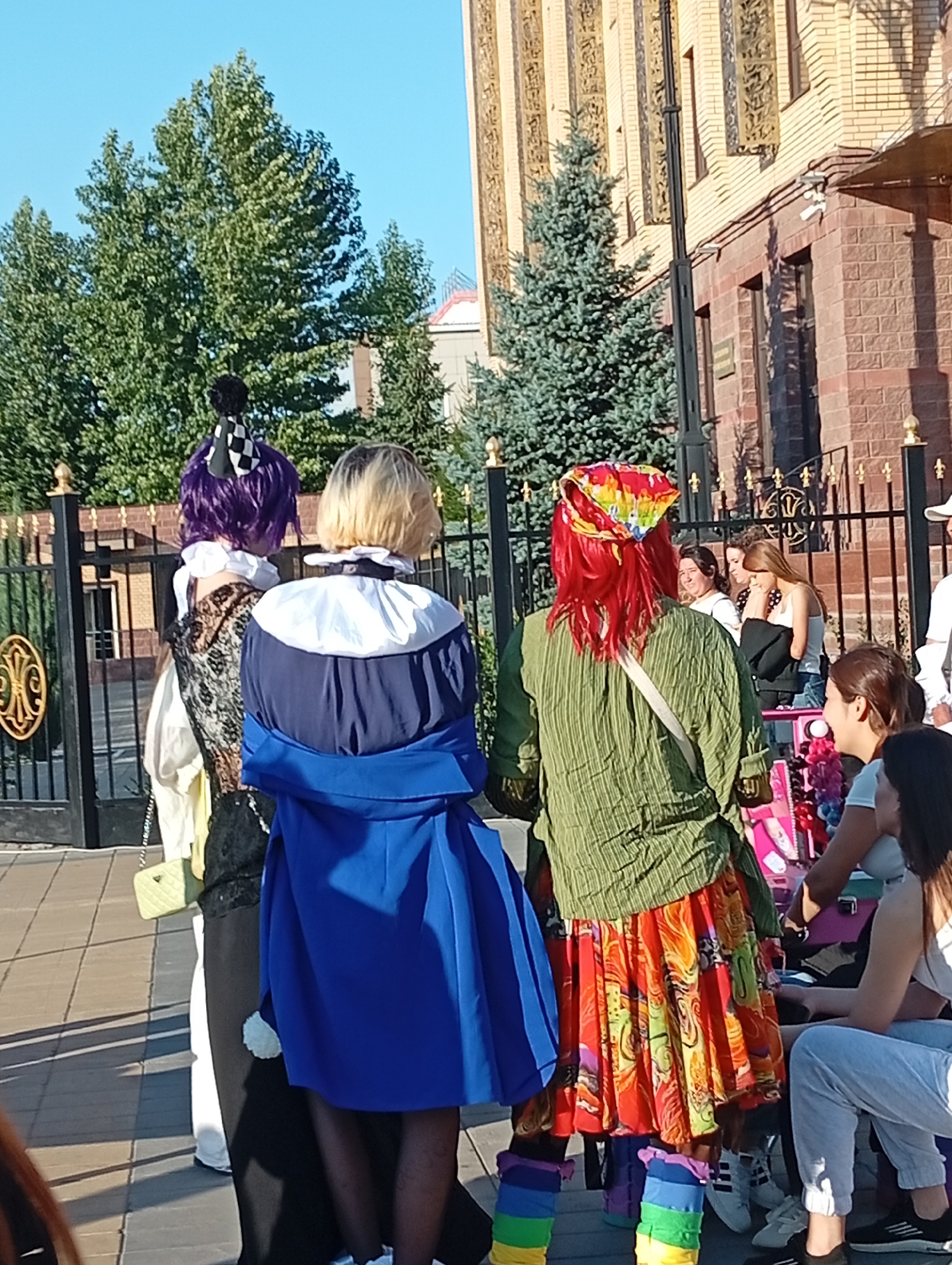An outdoor, medium shot shows a group of people in costumes standing on a paved area in front of a building and a fence. The building, made of brick and ornate stone, is on the right side of the frame. A black metal fence, topped with gold decorative elements, runs across the middle ground. Behind the fence are trees, including a tall, lush green deciduous tree on the left and a more slender, blue-green evergreen tree behind it. The sky is clear and blue.
In the foreground, three individuals are dressed in elaborate costumes. The person on the far left has purple hair and wears a dark, ornate patterned vest over a white shirt, with a checkered black and white hair accessory. To their right, another person with blonde hair wears a blue and white outfit with a ruffled collar. The person in the center has bright red hair and wears a colorful patterned headscarf, a green crinkled tunic, and a vibrant, multi-colored skirt. They are also wearing brightly colored leg warmers.
Further back, behind the fence, other individuals can be seen. A few women are looking towards the costumed people. One woman with dark hair is visible on the left, and two women with lighter hair are to her right. On the far right, a person in a white shirt and cap is partially visible. A table with various items, possibly merchandise, is set up to the right of the costumed individuals. The ground is paved with rectangular stones.