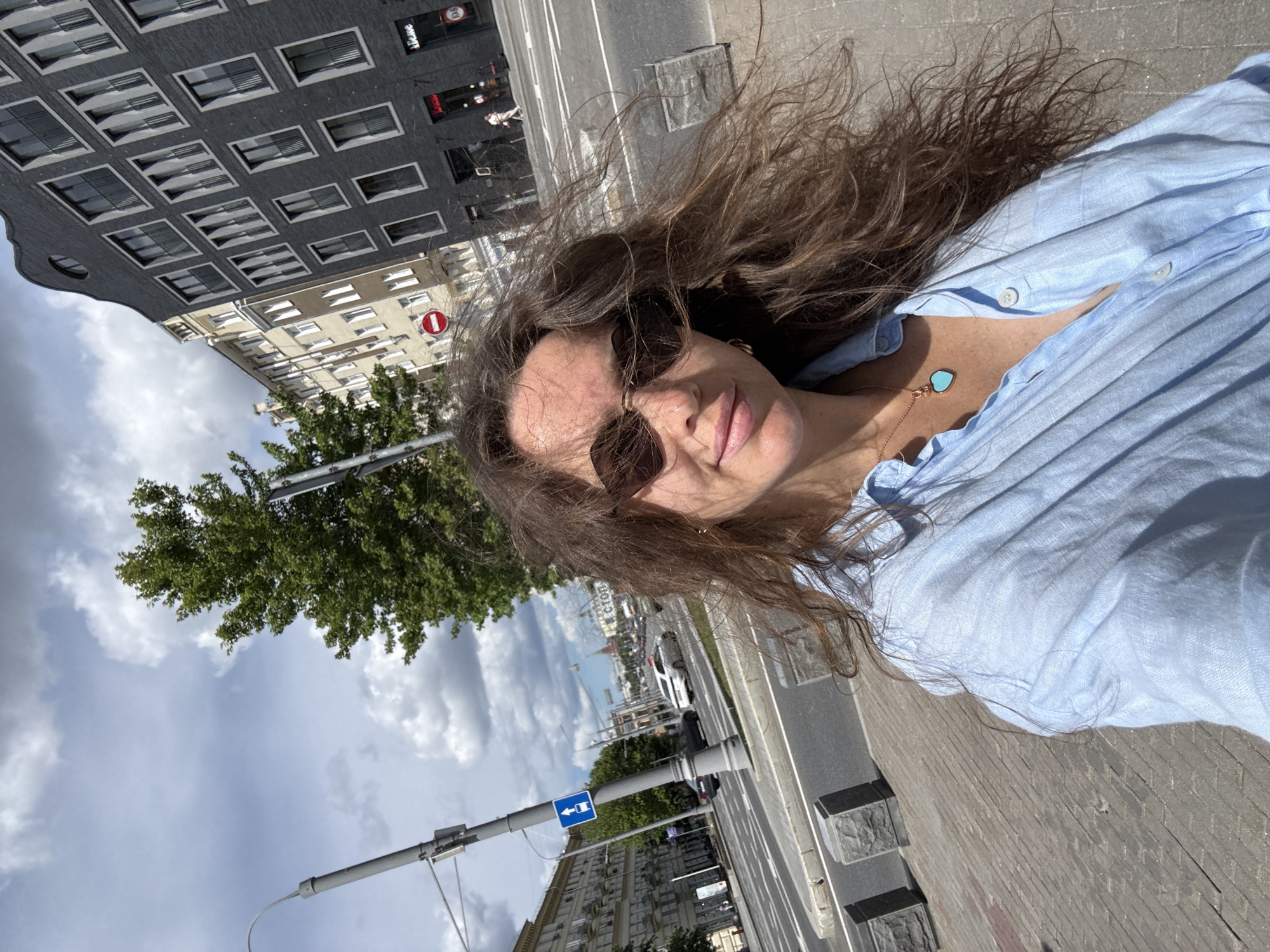 A woman with long, wavy brown hair and sunglasses takes a selfie outdoors on a sunny day in Moscow, Russia. She is wearing a light blue, unbuttoned collared shirt and a heart-shaped turquoise pendant necklace. In the background, a busy street with cars and pedestrians is visible, along with buildings and trees. The architecture suggests an urban setting.