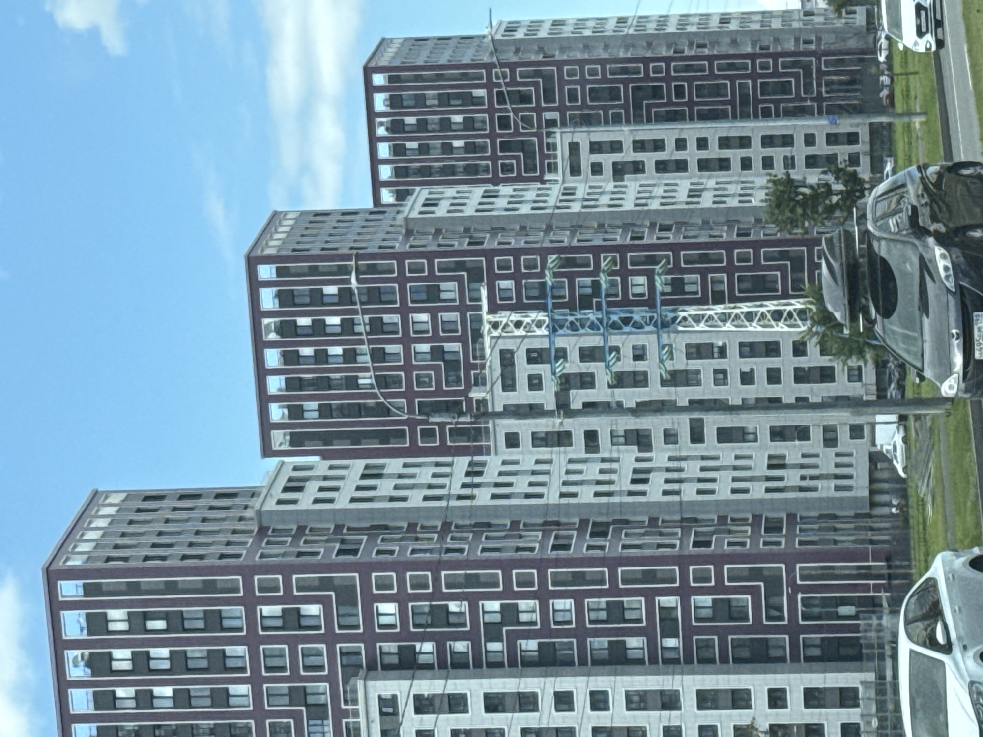 The image depicts a street scene in Saint Petersburg, Russia, featuring several modern, high-rise apartment buildings with distinctive architectural patterns. The buildings are predominantly gray and purple, with white geometric framing around the windows. In the foreground, there are several cars driving on a road, including a black SUV with a roof box and a white sedan. The sky is blue with scattered clouds. The scene appears to be a residential area with new construction.