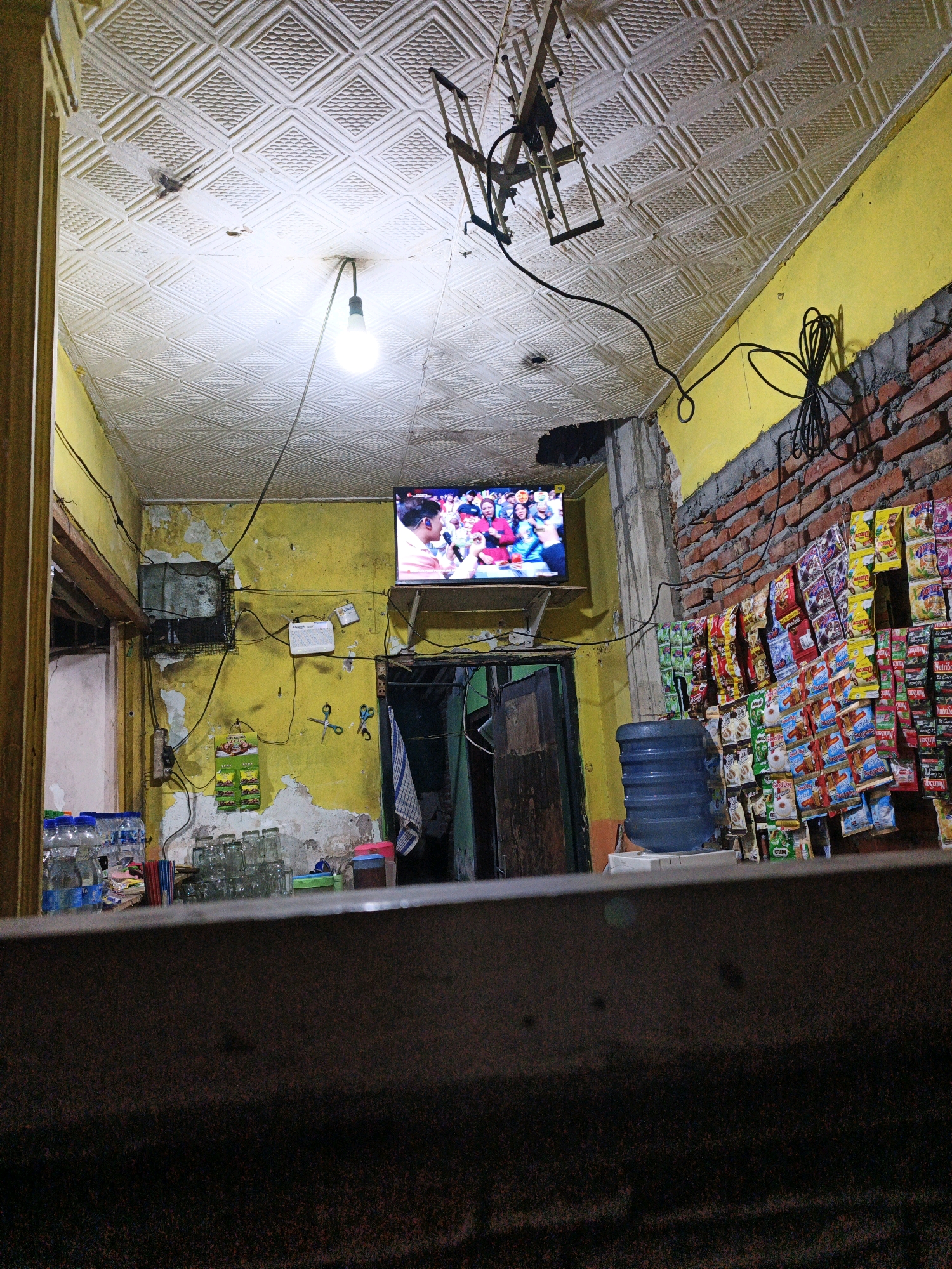 The image depicts the interior of a small shop or warung, likely in Ketegan, Indonesia, characterized by its rustic and somewhat cluttered appearance. The walls are painted a peeling yellow, and a section of exposed brick is visible on the right. The ceiling is textured, resembling embossed tiles.
Dominating the upper portion of the frame is a small television screen displaying a colorful image, possibly a sports event or a broadcast with multiple people. Below the television, the wall is adorned with various items: a white router, two small packets, a pair of blue scissors, and a wall-mounted electrical box. A prominent water dispenser with a blue tank sits to the right of the television, next to a stack of glass cups.
The shelves or display racks on the right wall are filled with numerous small, individually packaged food or beverage items, commonly found in such small Indonesian establishments. A single light bulb hangs from the ceiling, illuminating the scene. In the foreground, a dark, blurred surface suggests the presence of a counter or table. The overall impression is of a functional, lived-in space that serves as a local convenience store.
