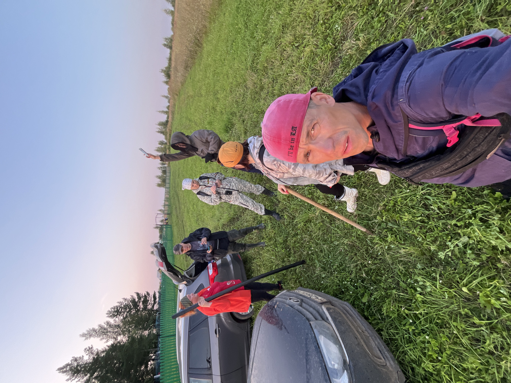 The image depicts a group of six individuals in an outdoor, grassy environment with two cars parked nearby. The scene appears to be located near the village of Derevnya Perebor, Russia, suggested by the overall setting of rural landscape and the presence of cars, implying a potential outing or activity.
In the foreground, a man wearing a pink baseball cap and a blue hooded jacket is taking a selfie, holding the camera with his left hand. He has a black fanny pack worn across his chest. His cap has text on it that reads "I'LL BE IN LA."
Behind him, to his right, a person in a camouflage-patterned outfit and a yellow beanie is holding a wooden pole. To the left of this individual, another person in a similar camouflage outfit and a white bandana is looking at their phone, which is held in their left hand.
Further back and to the left, a woman in a bright orange jacket and a patterned headband is holding a long, dark pole vertically with both hands. She is standing next to the open rear of a dark gray car.
Behind the woman in orange, a man in a dark jacket and a cap is standing near the open trunk of the gray car, holding a phone in his right hand.
To the right of the man in the dark jacket, a person wearing a dark hooded jacket with the hood up is holding a phone or similar object aloft in their right hand, seemingly pointing it towards the sky.
The environment consists of lush green grass with some dry, taller grass in the background. Trees are visible on the left side of the frame, and a wooden fence runs along the edge of the property. The sky is clear and shows a gradient of light blue and soft pink hues, indicative of early morning or late evening light. One of the cars is a dark gray SUV, and the other is a dark-colored car. The license plate of the dark-colored car in the foreground is partially visible.