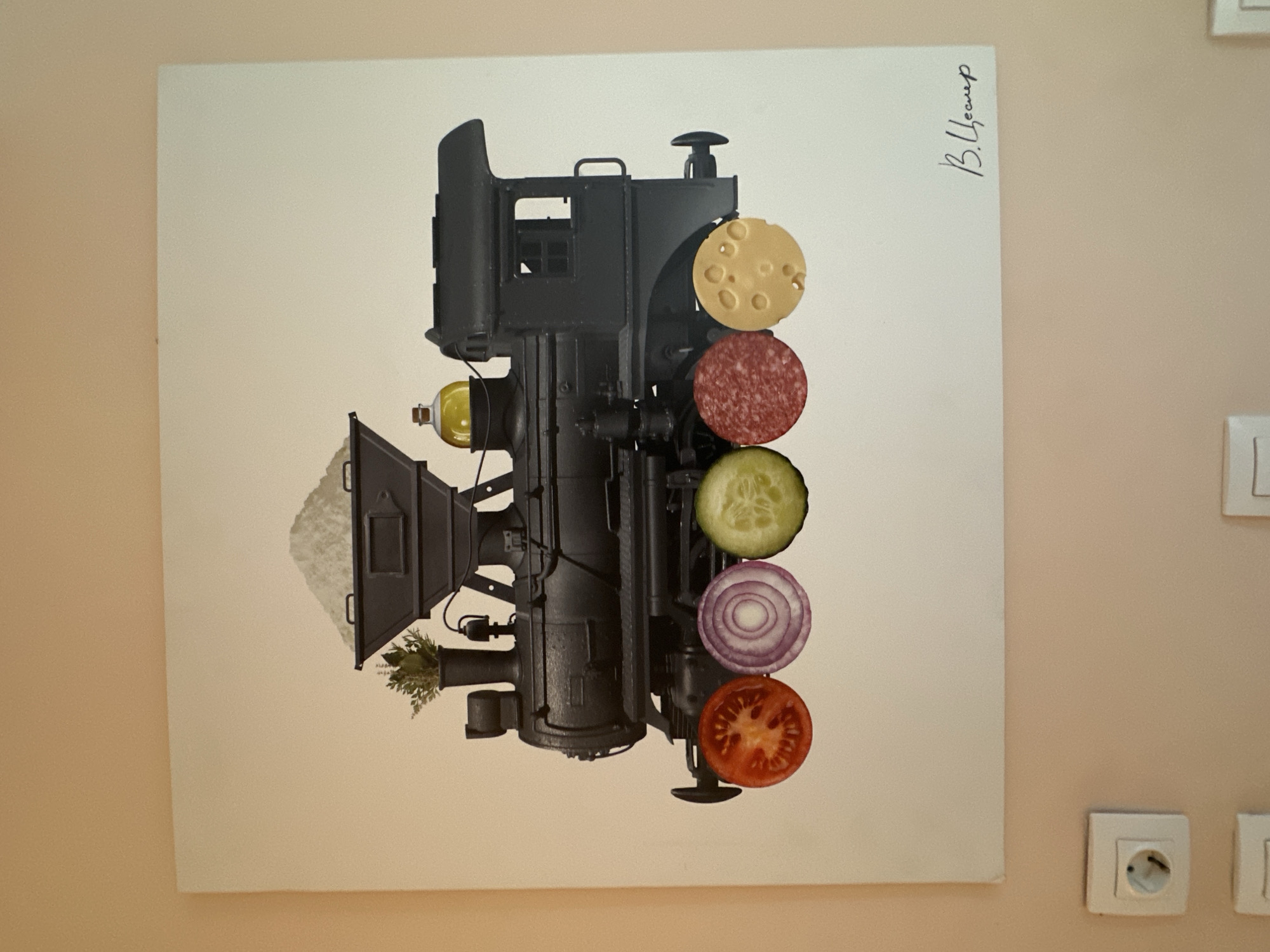 The image shows a painting of a black steam locomotive. The front of the train is decorated with slices of food: a tomato, an onion, a cucumber, salami, and cheese. The top of the train appears to be filled with salt, and there is a bottle of olive oil on the side. The painting is signed in the lower right corner. Below the painting, on a light pink wall, are two electrical outlets. The scene is located indoors, with the wall and electrical fixtures suggesting a residential or public building. There are no people visible in the image. The specific location in the city of Minsk, Belarus, cannot be determined from the visual information provided.
