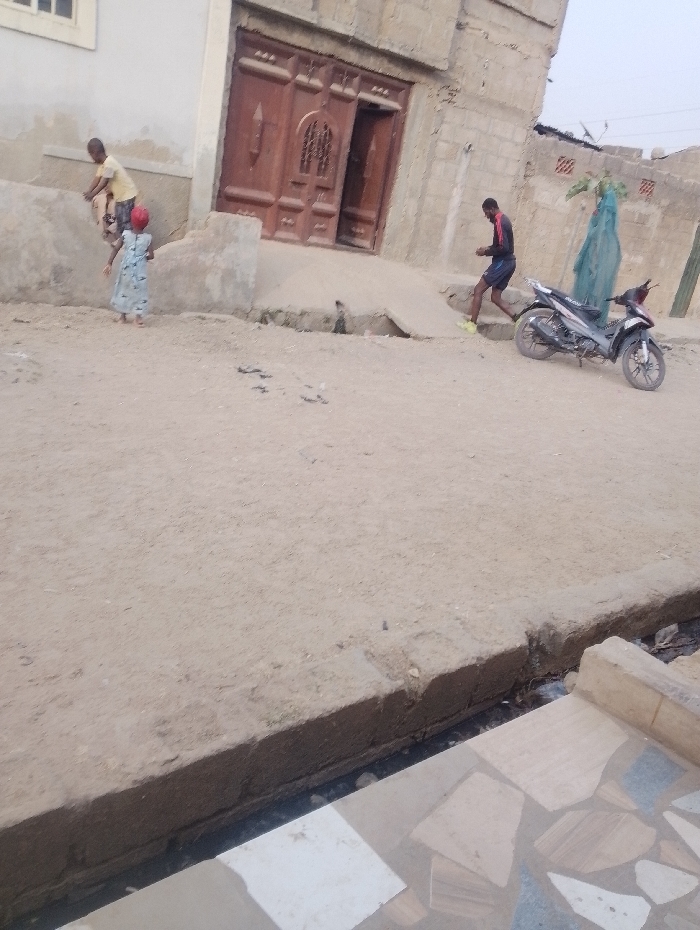 A street scene shows two children near a building's entrance, with one child on an elevated step. A man walks near a parked motorcycle. The buildings are constructed of neutral-toned materials, and the street is covered in a sandy dirt. A drainage area is visible at the bottom of the frame, with a decorated floor surface visible near its edge. The sky appears overcast.