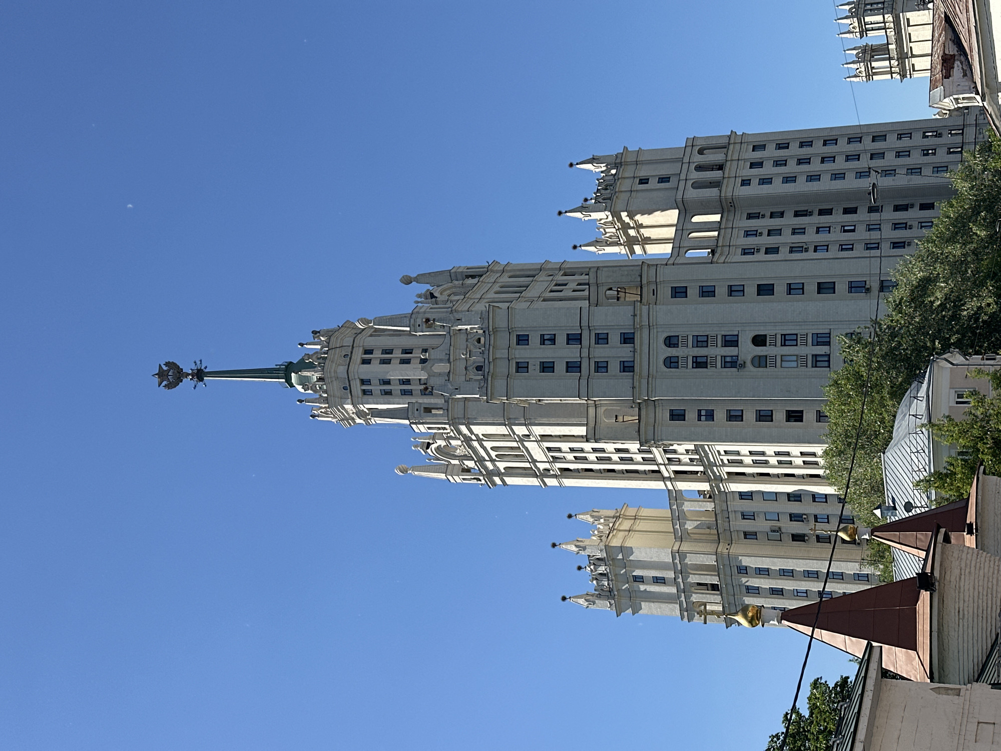 This detailed image depicts the Kotelnicheskaya Embankment Building, one of Moscow's "Seven Sisters" Stalinist skyscrapers, located in the Tagansky District of Moscow, Russia.
The primary subject is the massive, light-colored (off-white/cream) multi-tiered building, characterized by its ornate Stalinist Empire architectural style. A prominent central tower rises, topped by a dark green spire culminating in a golden five-pointed star. Numerous rectangular, dark-framed windows are visible across its facade. Various smaller towers, spires, and decorative elements adorn the building's many sections.
The sky is uniformly clear and bright blue, occupying the upper two-thirds of the frame. A faint, thin crescent moon is discernible in the upper right quadrant of the sky.
In the lower portion of the image, green deciduous trees are visible on the left and center. Parts of other buildings are present in the foreground: on the lower left, sections of structures with reddish-brown conical roofs, some featuring small golden domes topped with Orthodox crosses, and light-colored walls are seen. Dark power lines extend horizontally across the mid-ground. On the far right, a section of another light-colored building with decorative finials is partially visible. No people or discernible actions are present in the scene. The lighting suggests a bright, sunny day, with shadows indicating the sun is positioned to the right of the main building.