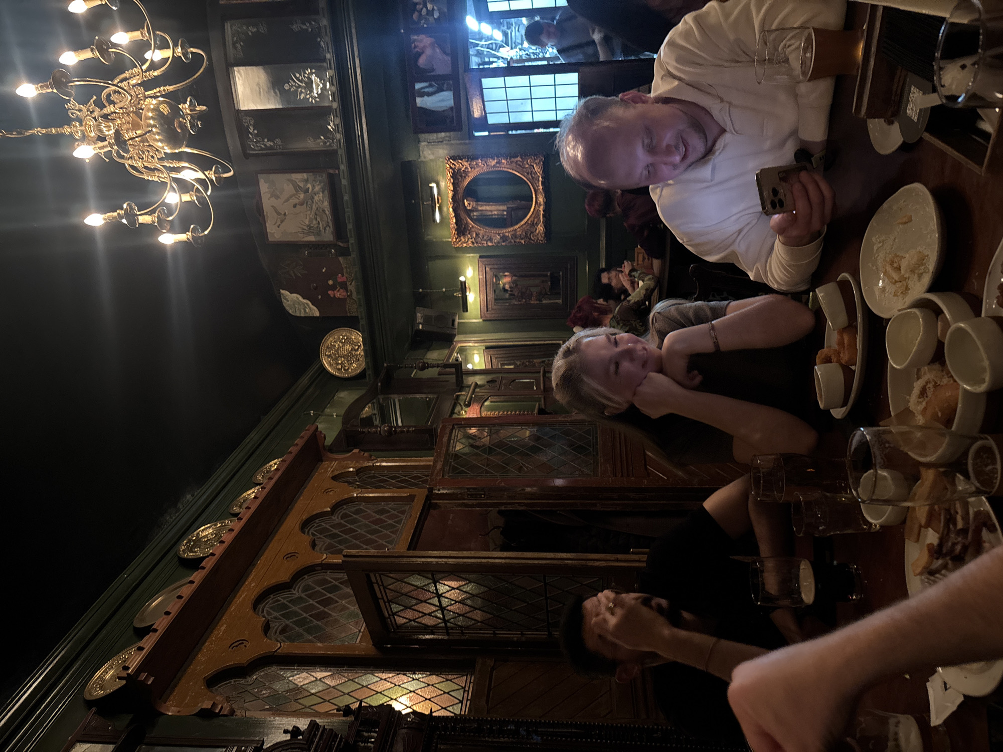 Here is a detailed description of the image:
**Setting:** The scene appears to be inside a dimly lit pub or restaurant with ornate, old-world décor. Details include a large, ornate chandelier, dark green walls adorned with framed pictures and decorative plates, and woodwork with stained glass panels.
**People:** There are multiple people visible. The composition focuses on a group seated at a table. A man in a white shirt looks down at his phone, another woman with blonde hair rests her head on her hands. Another dark haired person can only be seen in the lower section of the image.
**Objects:** The table is covered with drinks and remnants of food. There are beer glasses, plates with crumbs, empty mugs, and what appears to be a phone.
**Actions:** The man is using his phone, the woman seems to be relaxing at the table. The rest of the group are presumably engaged in conversation.
**Interactions:** The photo captures a casual social gathering. The composition hints at a relaxed and potentially long-standing interaction among the people at the table.
**Environment:** The dim lighting and the ornate details suggest a setting with a specific, possibly historic or traditional, atmosphere.