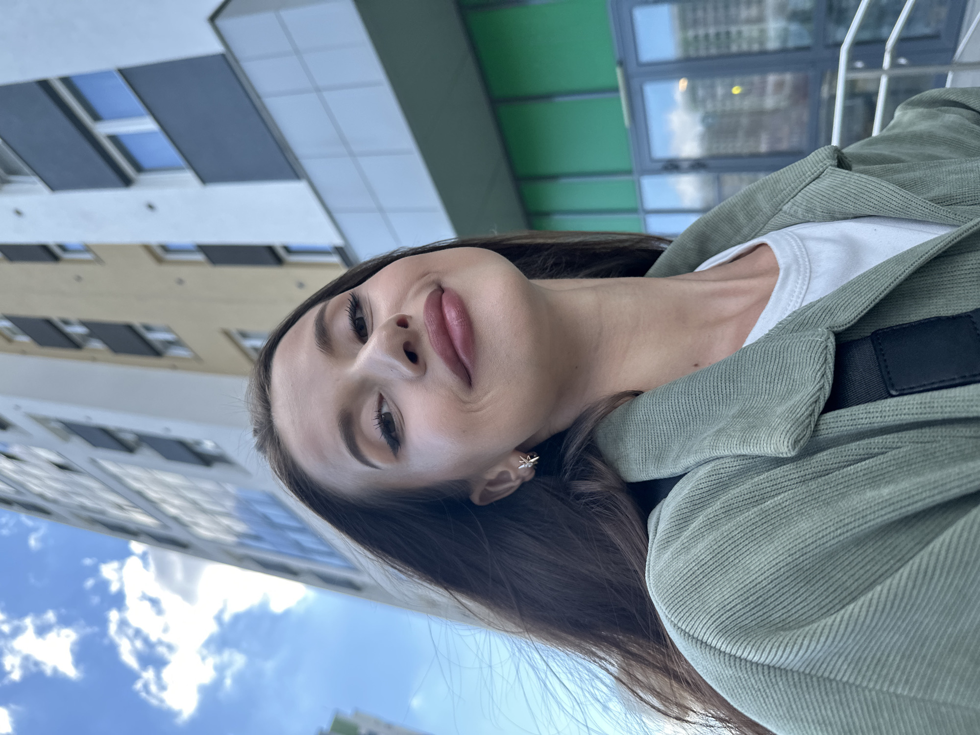 In Yekaterinburg, Russia, a young woman with long, dark hair and a light complexion is captured in a close-up, low-angle selfie. She is wearing a white t-shirt and a light green corduroy jacket, accessorized with a delicate star-shaped earring. Her facial expression is a gentle smile, with her head tilted slightly upwards towards the camera. Behind her, a modern residential building with a beige and white facade dominates the background, its windows reflecting the blue sky and scattered clouds. The architecture features dark gray balconies and green-tinted glass on the lower levels. The overall scene is set against a bright, daytime sky.