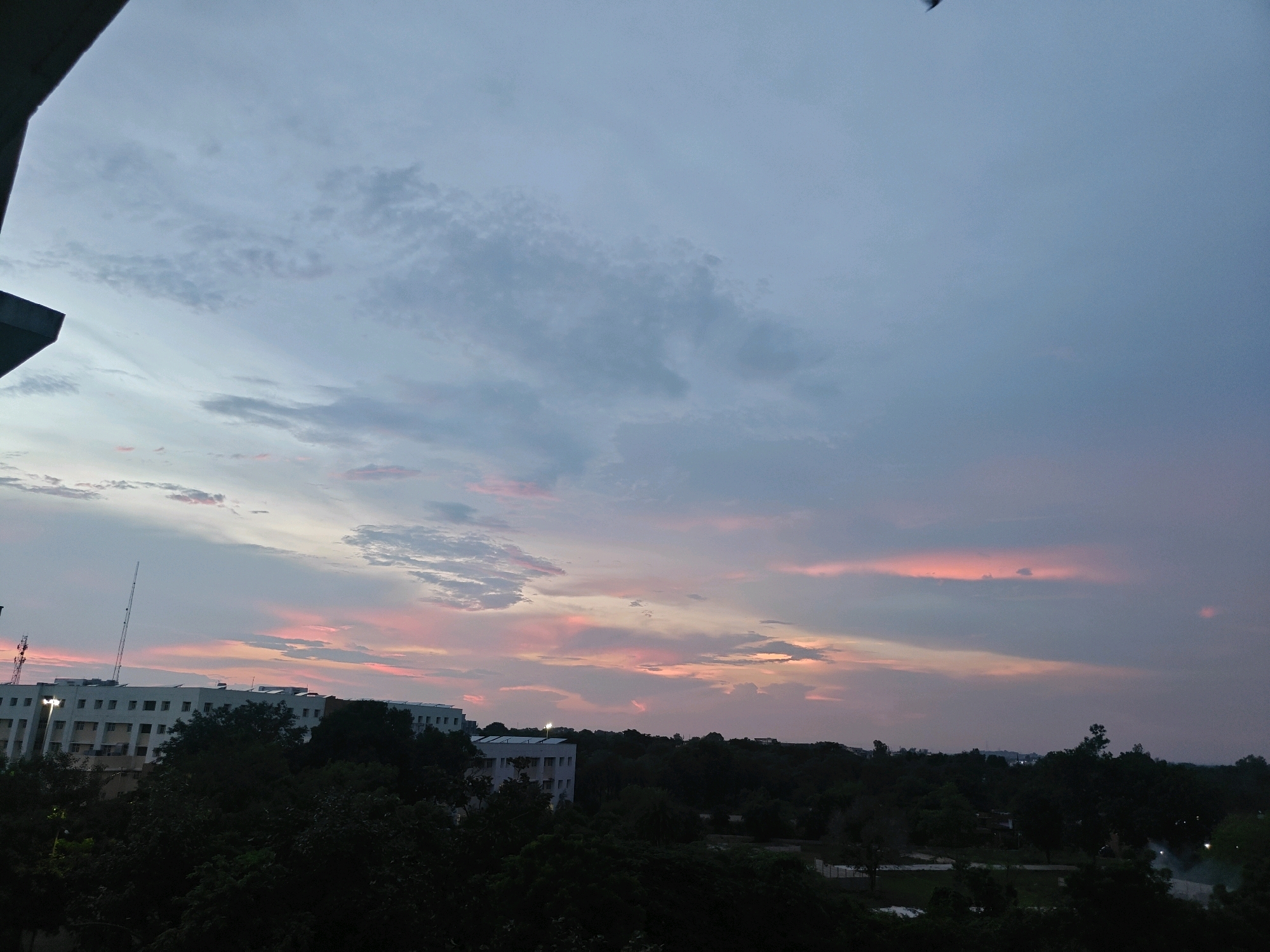 An image captures a sunset over Datia, India, showing a twilight sky with streaks of pink and orange clouds against a darkening blue expanse. In the foreground, silhouetted trees obscure the lower part of the scene. A long, multi-story building with a flat roof and numerous windows is visible on the left, partially covered by dark foliage. A tall, slender antenna rises from behind this building. Further in the background, more trees and the tops of other structures can be discerned against the dimming sky. The overall impression is of a peaceful, late evening in an urban or semi-urban setting.