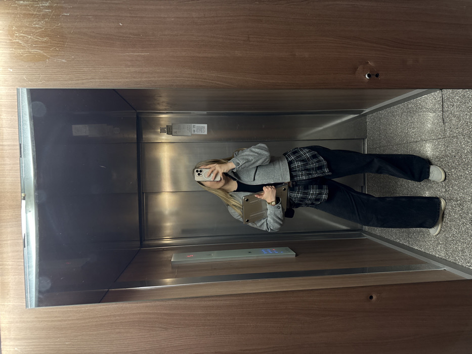 An elevator located within the city of Budva, Montenegro, is depicted. The interior features dark wood paneling on the side walls and a brushed metallic back wall. A vertical elevator control panel displaying a red "5" is present on the left wall. The floor is composed of a light-colored speckled material. One individual, appearing female with long blonde hair, is standing in the center of the elevator, facing a reflective surface on the inner door. The individual is actively taking a photograph using a light-colored smartphone, which is held in front of their face. They are wearing a black short-sleeved top, a gray and black plaid shirt tied around the waist, a cropped gray jacket with button details, dark wide-leg jeans, and light gray sneakers. A silver or metallic-colored laptop or tablet, enclosed in a clear case, is held in the individual's left arm. A watch is visible on their left wrist, and a dark strap, possibly from a bag, is visible over their left shoulder.