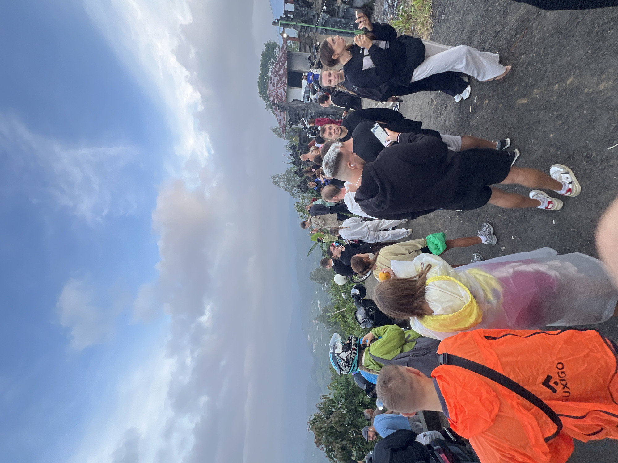 The image shows a crowd of people gathered on a paved surface in what appears to be an elevated location. The location is the city of Kintamani in Indonesia. Many individuals are wearing casual clothing and some are wearing rain gear like ponchos. Some people are using their mobile phones. A mountain range and lush vegetation can be seen in the background. A cloudy sky is overhead. Some motorcycles are parked nearby.