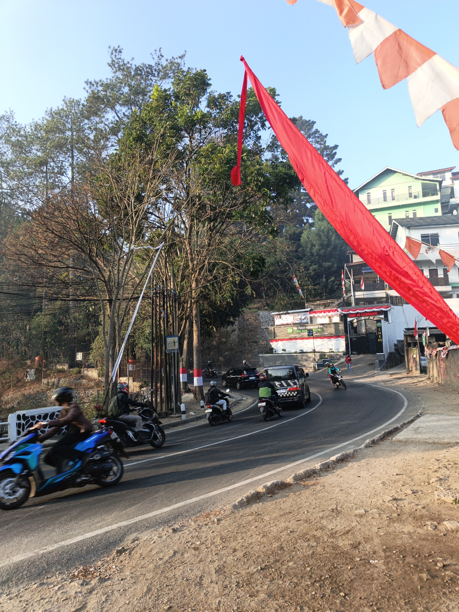 The image depicts a winding road in Lembang, Indonesia, lined with trees and buildings. Several motorcycles and a black SUV are visible on the road, suggesting a typical day of traffic. Large red and white flags are prominently displayed, indicating a festive or nationalistic occasion. Buildings in the background, possibly schools or businesses, are decorated with smaller flags. The environment appears to be a hilly or mountainous region, given the dense vegetation and the upward slope of the road. The lighting suggests it is either early morning or late afternoon.