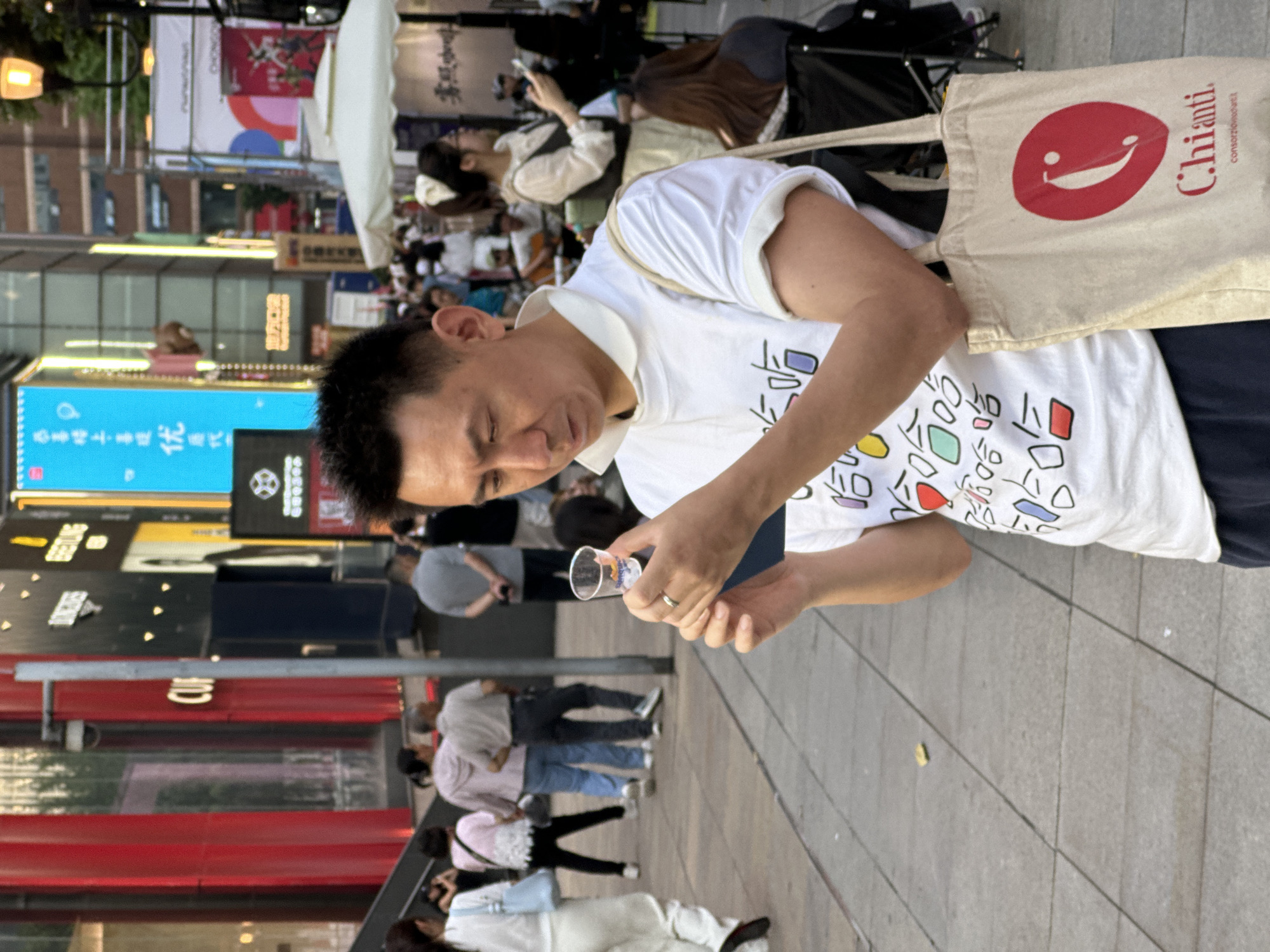 The image, rotated 90 degrees clockwise, depicts an outdoor urban street scene in Yuzhong District, China, likely during late afternoon or early evening.
The main subject is a man, appearing to be in his 30s or 40s, with short dark hair. He wears a white short-sleeved polo shirt featuring colorful, abstract-style Chinese characters or symbols, and dark-colored pants. A beige canvas tote bag is slung over his left shoulder, adorned with a large red smiling face logo and the word "Chianti" in red script, with smaller text "consorzio del Chianti" below it. The man is looking downwards with a focused expression, holding a small, clear plastic cup in his right hand and a dark-colored object, possibly a smartphone, in his left. A silver-colored ring is visible on his left ring finger.
In the midground and background, numerous other people are visible, some walking and others seemingly seated at outdoor establishments. The street is lined with multi-story buildings, featuring large digital screens and storefronts. One prominent blue digital screen in the background displays Chinese characters, including "优" (yōu - meaning 'excellent' or 'superior'), indicating a commercial advertisement. Another street lamp with an orange glow is visible in the upper left corner of the frame. The ground is paved with light-colored tiles, characteristic of a pedestrian area in a bustling commercial district.