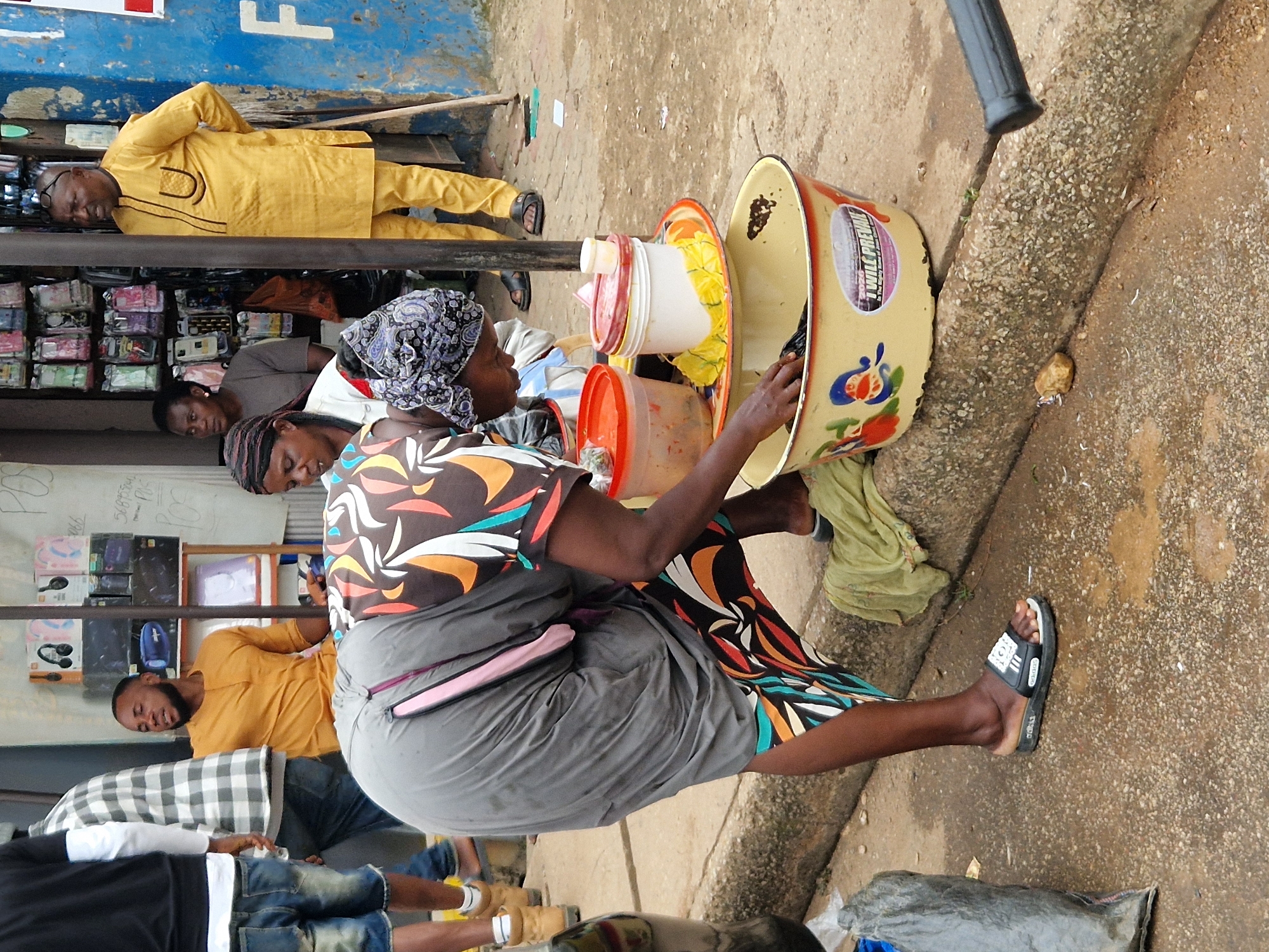 The image depicts an outdoor scene, likely a marketplace, with several individuals present. In the foreground, a person wearing a gray dress and a colorful patterned skirt is bending over a yellow basin, reaching into it with their left hand. In front of this person are two stacked plastic containers, one red and one white, on a yellow tray. To the right, a man in a yellow traditional outfit stands with his hands behind his back, looking towards the left. Behind him, a stall displays various items, including mobile phone cases. To the far left, a person in a black t-shirt and denim shorts is partially visible. The ground is wet and appears to be paved with concrete and gravel. A green cloth is draped over a concrete curb to the right of the person in the foreground. The overall environment suggests a humid, possibly tropical climate.