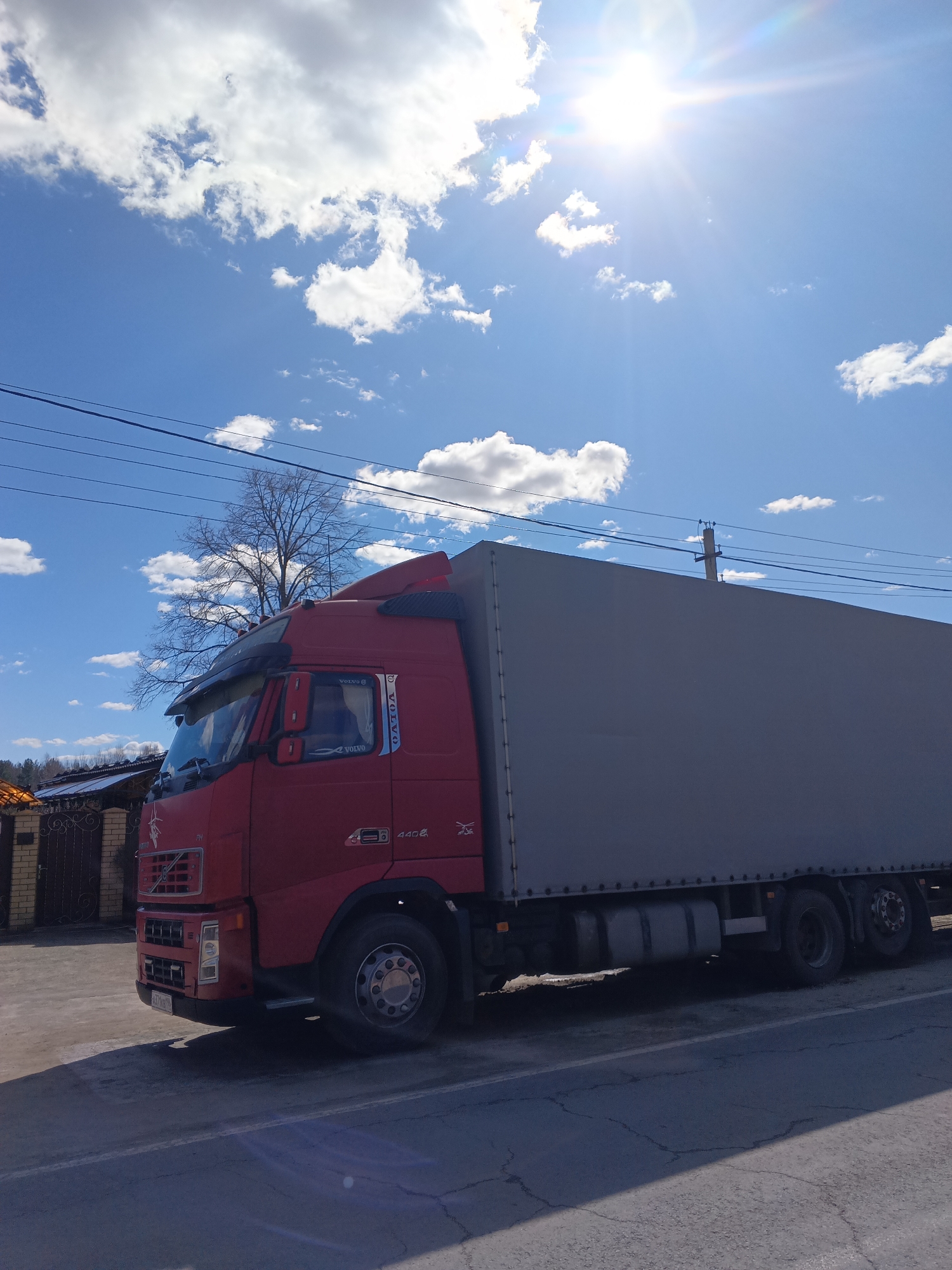 A red Volvo FH series truck is parked on the side of a cracked asphalt road in Красный Маяк, Russia. The truck has a grey, boxy cargo trailer attached. A short brick wall is visible in the background. A bare tree stands to the left of the truck's cab. Utility lines are strung overhead against a blue sky with scattered white clouds and a bright sun. The sun's glare dominates the upper portion of the image.