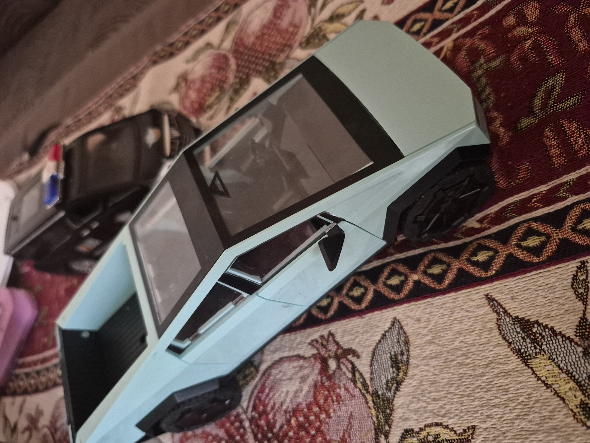 The image captures a light blue, futuristic-looking toy truck, possibly inspired by the Tesla Cybertruck, resting diagonally on a richly patterned textile. The toy features distinct angular lines, black windows, and black wheel wells, with the front part prominently visible in the upper right.
In the blurred background, behind and to the left of the main toy, another dark object is partially visible, showing small red and blue elements, possibly part of another toy or an electronic device.
The surface beneath the toy is a decorative rug or blanket, characterized by traditional patterns in shades of red, beige, black, and yellow. Visible motifs include stylized pomegranates and leaves, along with geometric borders. These traditional patterns could suggest a cultural context common in regions like Armenia, aligning with the provided location of Yerevan.
The scene appears to be an indoor setting, illuminated by ambient light. The photograph is taken from a slightly elevated perspective, looking down, with the objects arranged at an angle. No discernible activity is taking place, and no text is visible in the image.