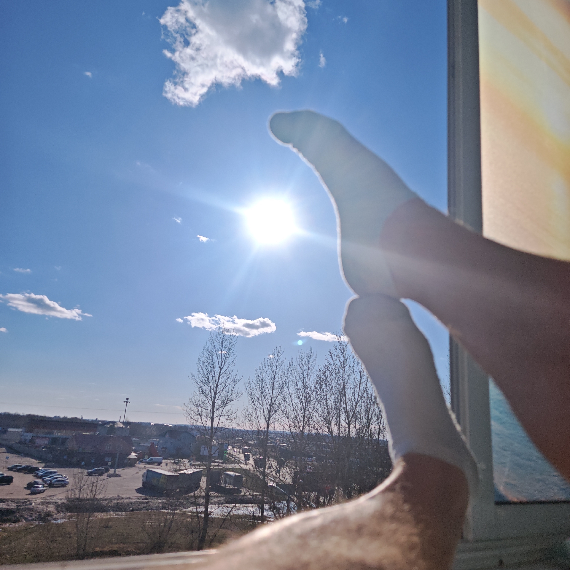 The image shows a view from a window, with feet and legs wearing white socks in the foreground, partially obscuring the view outside. The sky is bright blue with scattered white clouds, and the sun is shining prominently. Below the sky, there is a view of a city. Bare trees are visible in the middle ground. In the distance, there are buildings and a parking lot filled with cars. Based on the buildings and landscape visible, the location is likely the city of Rostov, Russia. The image is taken from inside a building, looking out a window. The legs are crossed at the ankles and in the lower right there's the edge of the window.