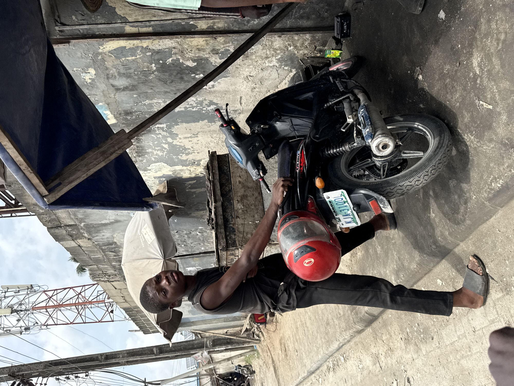 The image, taken in Lekki, Nigeria, shows a young man standing outdoors next to a black motorcycle under a makeshift shelter. He is dark-skinned, wearing a black tank top, black pants, and black flip-flops. He holds the motorcycle's handlebar with one hand, while a red helmet hangs from the bike. His expression is neutral as he looks towards the camera.
The setting is an urban, somewhat dilapidated environment. Behind the man and motorcycle, a wall with peeling light-colored paint is visible, and the ground is cracked concrete. Overhead, a dark blue tarpaulin or fabric provides shelter, supported by wooden beams. A beige or khaki-colored umbrella is partially visible next to the man, possibly folded or used for shade.
In the background, under an overcast sky, a large red and white communication tower is prominent, with various electrical wires crisscrossing the frame. Partially visible hands or figures are at the top left, suggesting other people nearby.
Notable details include the motorcycle's rear license plate, which reads "LAGOS KJA-310QL". A black battery pack with "AUKU" visible on its label is on the ground near the motorcycle. The umbrella also has some visible text, possibly "BATTERY SAFE". The scene is captured during the daytime under an overcast sky, suggesting a typical day in a bustling street environment.