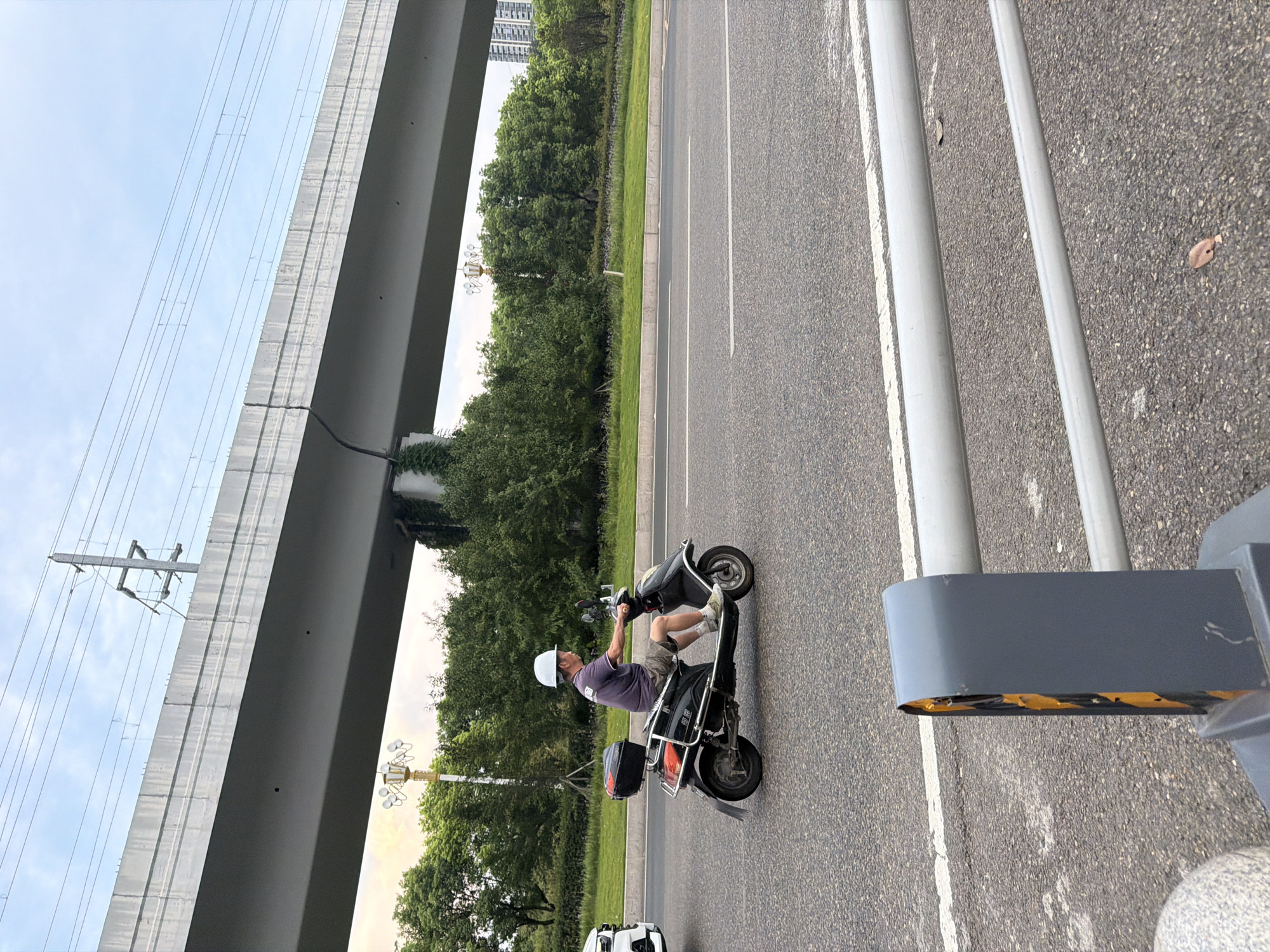 A man wearing a white helmet and a purple t-shirt rides a black electric scooter down a paved road in Jiangjin, China. The scooter has a red and black storage box attached to the rear. The man is wearing khaki shorts and white sneakers. The road has white lane markings and is bordered by a grassy median with trees. An elevated concrete structure, possibly a bridge or overpass, runs overhead, with power lines visible against a pale blue sky. In the background, tall buildings and more trees can be seen. A gray pole with yellow and black markings is in the foreground, part of a street barrier.