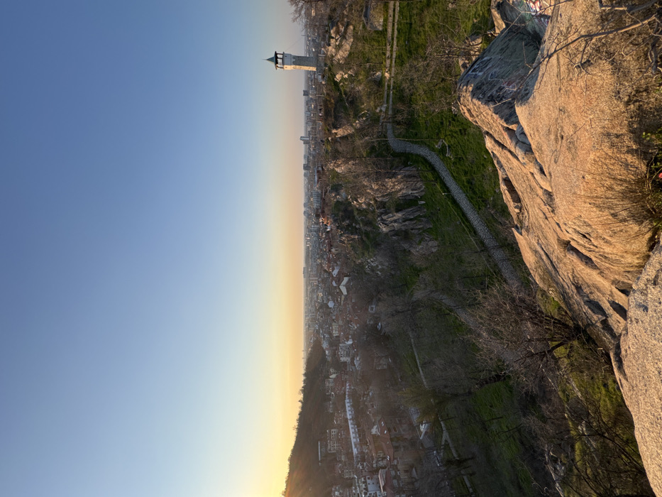 The image presents an elevated, wide-angle view overlooking the city of Plovdiv, Bulgaria, specifically captured from one of its hills, likely Nebet Tepe, looking towards Sahat Tepe (Clock Tower Hill) and the urban sprawl. The sky dominates the upper portion, exhibiting a clear gradient from light blue overhead to a warm yellow and orange at the horizon, indicating late afternoon or early evening light, with sunbeams visible on the far left.
In the immediate foreground, a rocky outcrop with rough, light brown and orange-hued stone formations occupies the lower right quadrant. Sparse, dry-looking vegetation, including bare branches and some green grass, grows among these rocks.
The midground features a winding, unpaved or cobblestone-like path that traverses a steep, grassy slope populated with numerous bare trees and shrubs. Rocky formations are interspersed throughout this terrain. To the right, a prominent, light-colored stone clock tower with a distinct green, pointed roof (the Sahat Tepe Clock Tower) stands atop a distant hill.
The background reveals a dense urban landscape stretching across the panorama. Numerous buildings with varying architectural styles and sizes, including residential structures with red-tiled roofs and taller, more modern constructions, fill the cityscape. A forested hill rises on the far left, partially illuminated by the setting sun. No individuals or specific actions are discernible within the scene.