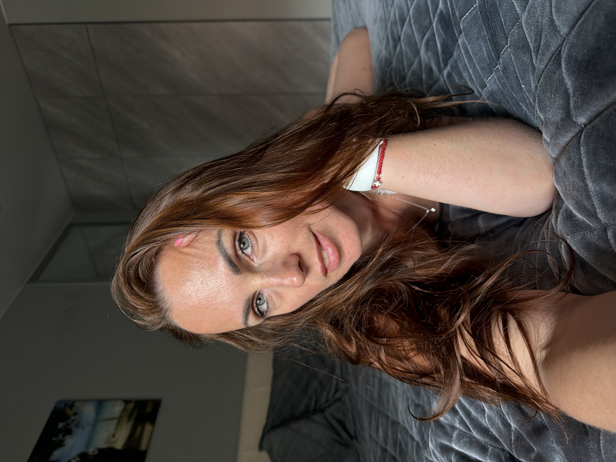 A woman with long, reddish-brown hair is lying on a gray, quilted bedspread, resting her head on her arm. She is looking towards the camera with blue eyes and a slight smile. A thin silver necklace with a small diamond pendant is visible around her neck. On her wrist, she wears a red string bracelet with a small white charm. A light-colored band is also visible around her wrist. She has a small pink mark on her forehead. The background shows a wall with a textured, gray paneling and a hint of a framed picture on the left. The overall scene appears to be indoors, possibly a bedroom, with soft lighting. There is no indication of the location being Birobidzhan, Russia, or any specific urban setting.