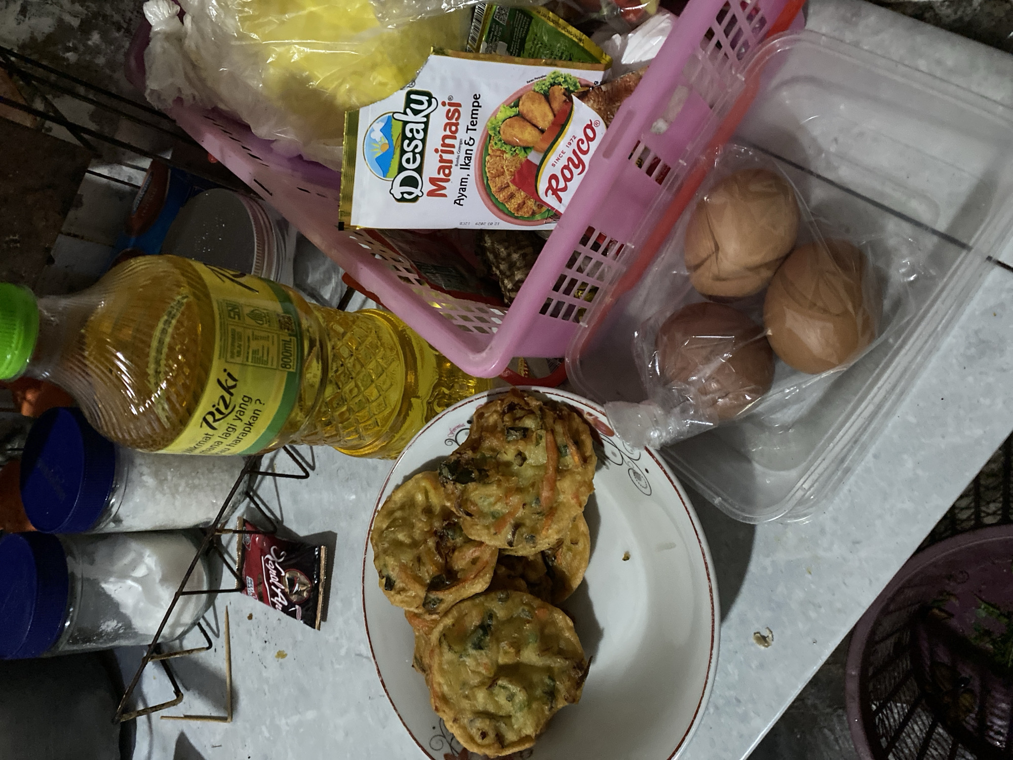 A close-up, eye-level shot captures a kitchen counter scene in Madiun, Indonesia. In the foreground, a white bowl holds several golden-brown fritters, possibly bakwan, with visible vegetable pieces. To the right, a pink plastic basket contains various packaged food items, including a sachet labeled "Desaku Marinasi" and a yellow plastic bag. A bottle of "Rizki" cooking oil stands tall in the background, beside two glass jars with blue lids, one filled with white granulated sugar and the other with a white powdery substance. A small, red foil packet, likely a seasoning or snack, rests on a metal rack. A clear plastic container holds three brown eggs. The surfaces appear to be a textured countertop, with a hint of a woven basket in the lower left corner. The lighting suggests an indoor setting, possibly a home kitchen.