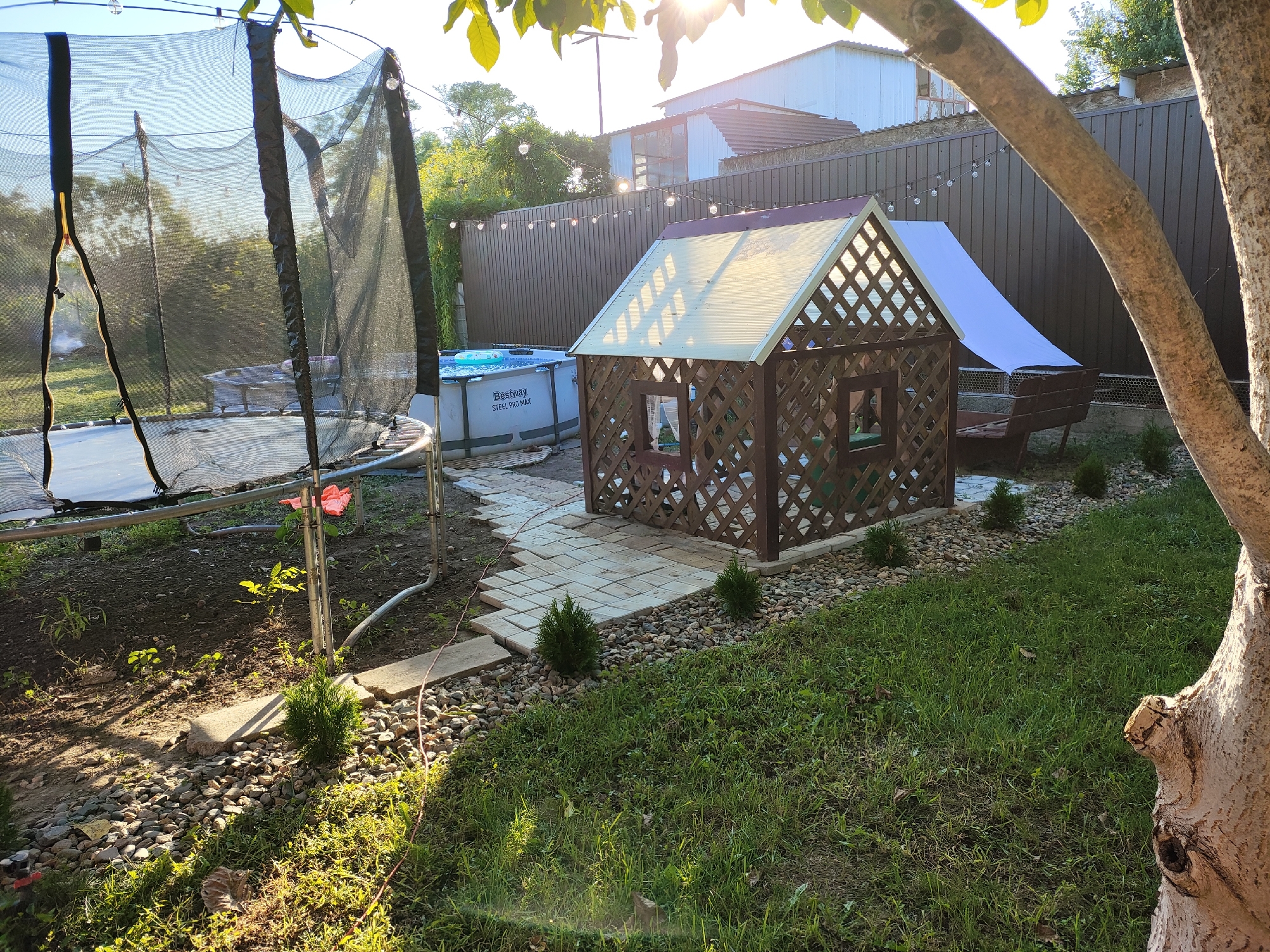 In the city of Medvyodovskaya, Russia, a backyard scene is captured on a sunny day. The foreground features a large, above-ground swimming pool with a visible "Bestway Steel Pro MAX" label. Adjacent to the pool is a wooden playhouse, intricately designed with a lattice pattern and a translucent, peaked roof. A stone pathway leads from the pool towards the playhouse. To the left, a trampoline with a black safety net is partially visible, its metal frame reflecting sunlight. The ground is a mix of dirt, gravel, and green grass. In the background, a dark brown wooden fence encloses the yard, and a string of decorative lights is visible above it. A large tree trunk, textured and aged, dominates the right side of the frame, partially obscuring the view. The overall environment suggests a residential backyard equipped for recreation.
