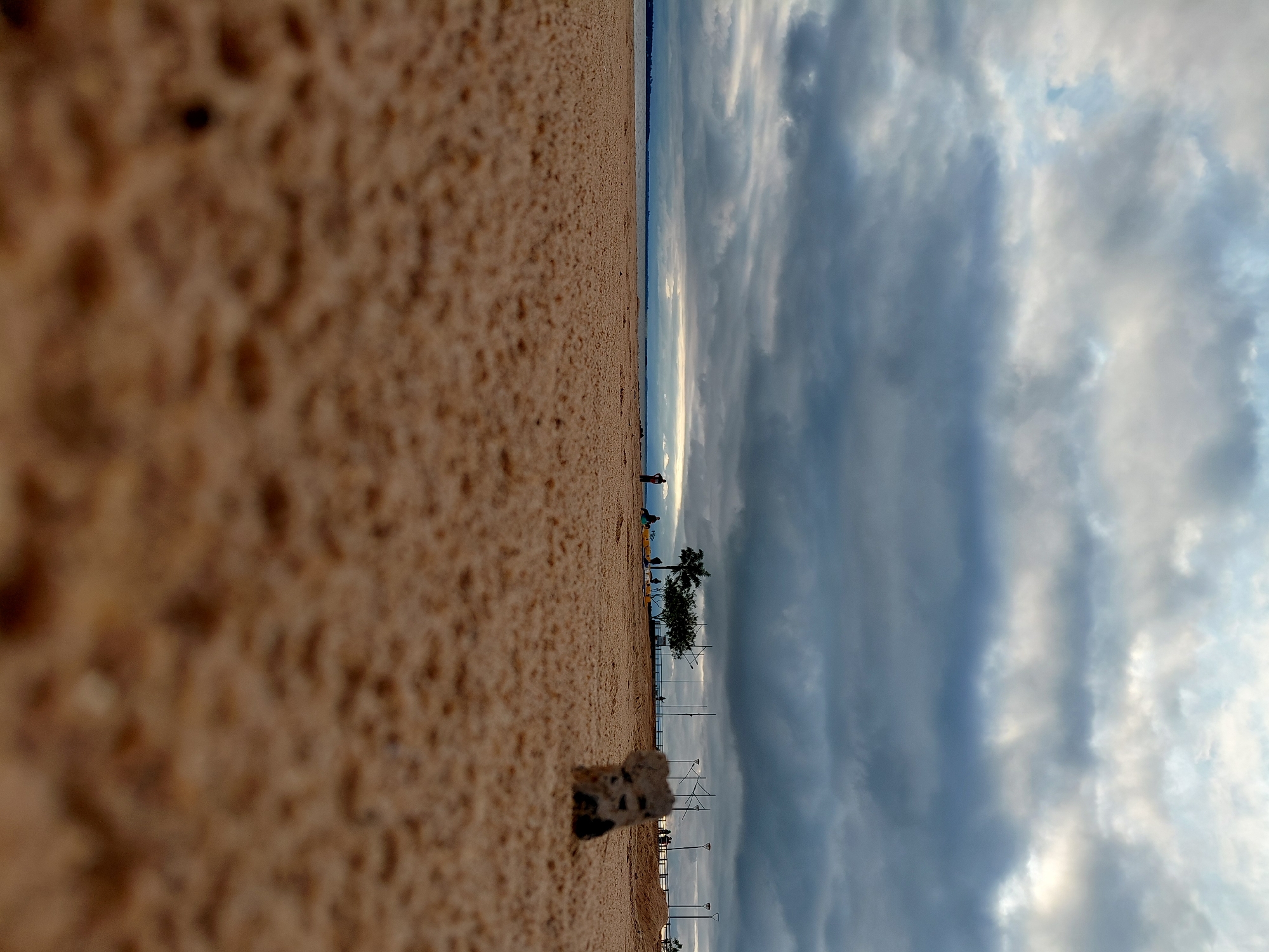 A low-angle, close-up shot focuses on the sandy shore of the Paraná River in Encarnación, Paraguay. The sand is the dominant feature of the foreground, with a small, dark, porous object in sharp focus on the right. Beyond the beach, the water stretches to the horizon, meeting a sky filled with dramatic, heavy clouds tinged with sunlight.
In the middle ground, a few people are scattered along the promenade, some standing, others appearing to be sitting. A lone palm tree and other foliage are visible, along with lampposts lining the walkway. The scene suggests a late afternoon or early evening setting, with the overcast sky contributing to a subdued atmosphere. The overall impression is one of a calm, perhaps slightly melancholic, end-of-day scene at the waterfront.