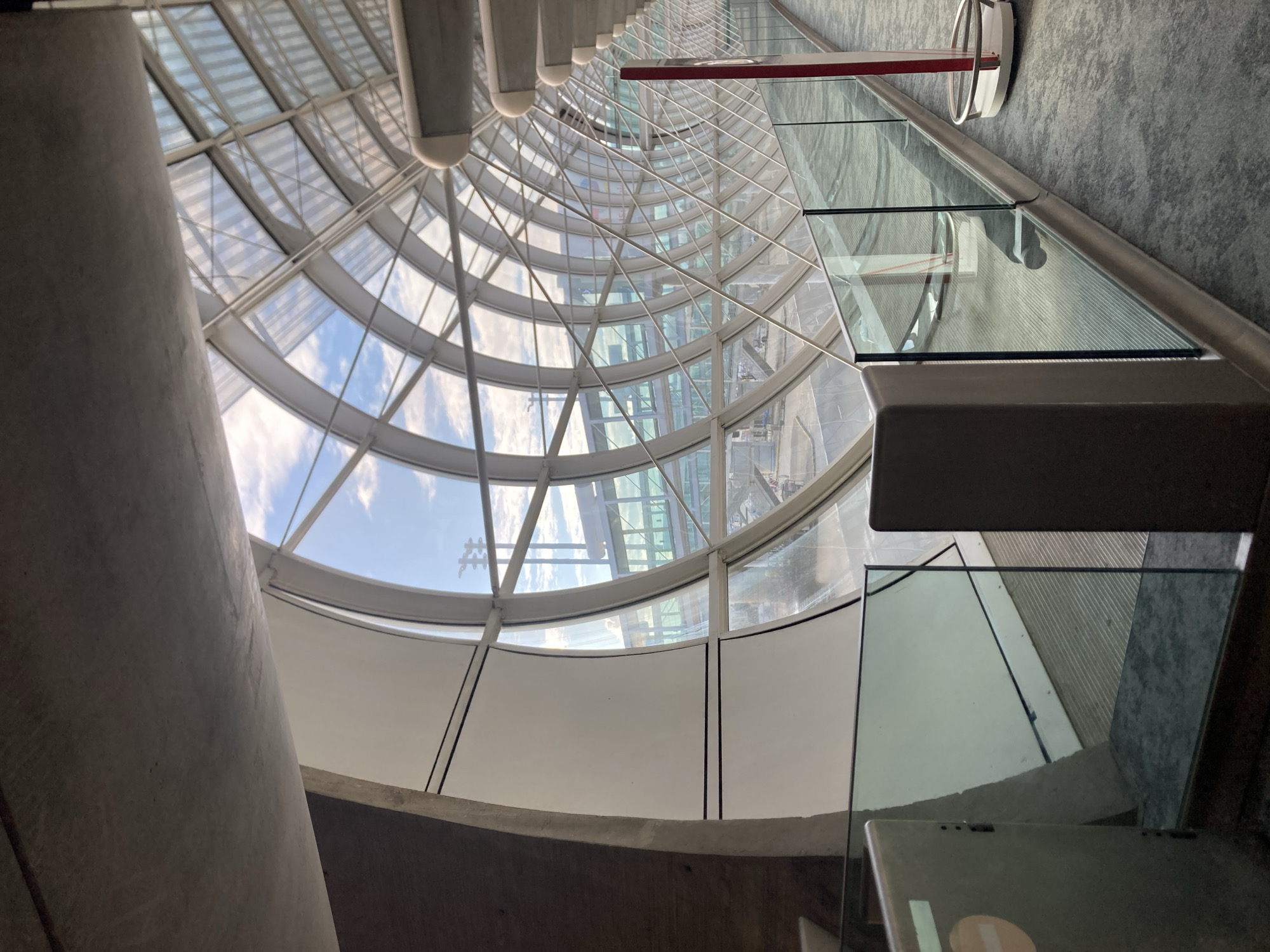 A low-angle, distorted perspective captures the interior of a cylindrical building in Le Mesnil-Amelot, France. The structure appears to be modern, featuring a curved glass facade supported by a white metal framework. The glass panels reveal glimpses of a partly cloudy sky. Sections of the facade are opaque, possibly made of a white material. A grey concrete column is visible in the foreground on the left. Parts of a glass-paneled staircase or walkway with a metal railing and grey floor are visible on the right. A red and white circular fixture is attached to the glass.
