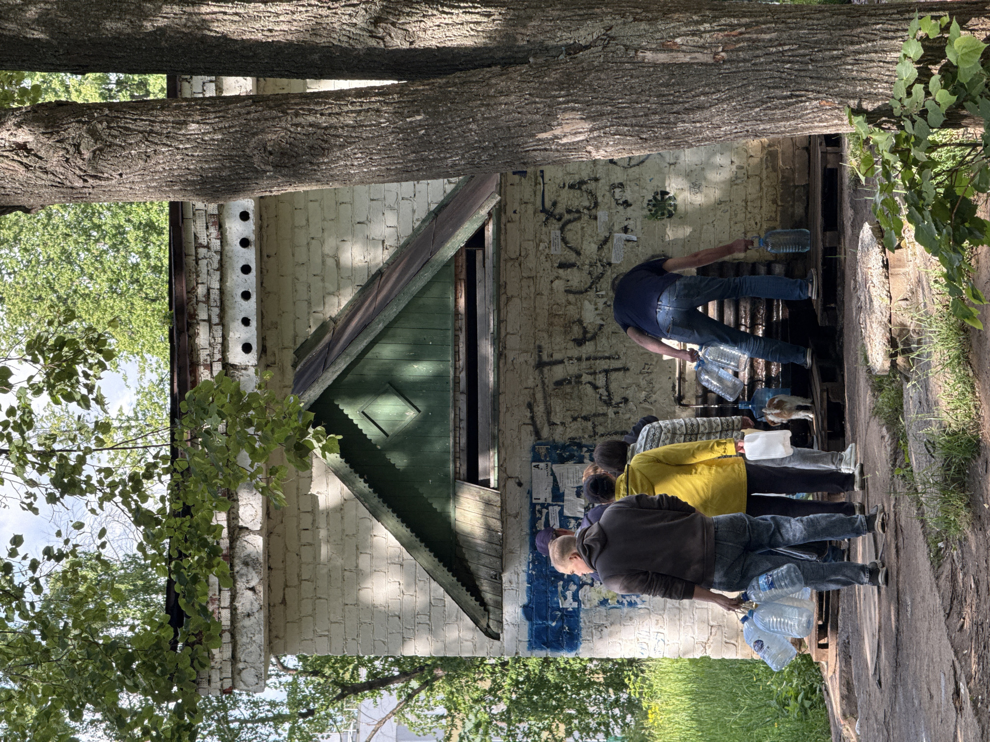 The image, taken in Chaikovsky, Russia, captures an outdoor scene where several people are collecting water from what appears to be a public or natural water source. The setting is bright and sunny, indicated by the distinct shadows cast by trees and structures.
In the foreground, two men are prominent. One man, dressed in a dark brown jacket and blue jeans, holds a collection of clear plastic water bottles by their handles. Slightly behind him, another person in a yellow jacket is partially visible, also carrying water containers. Further to the right, a man in a dark blue t-shirt and jeans is bent over a dark wooden structure, actively filling large plastic bottles with water. Numerous other empty and full plastic bottles, typically 5-liter size, are seen on the ground and on the wooden platform surrounding the water source. A small cat is visible near the feet of the person in the yellow jacket, possibly resting or drinking.
The backdrop features a dilapidated, white-painted brick building. Its walls are adorned with extensive dark graffiti, streaks of blue paint, and several faded pieces of paper or posters, one of which clearly displays a large black capital "A." A dark green, sloped roof or awning section with intricate wooden detailing extends from the building. Two prominent tree trunks with rough bark frame the left side of the image, while their branches with lush green foliage contribute to the natural, somewhat unkempt environment. The ground is a mix of dirt and patches of grass. The overall scene depicts a functional, everyday activity of water collection at a communal spot. The text on the posters and graffiti is largely illegible.