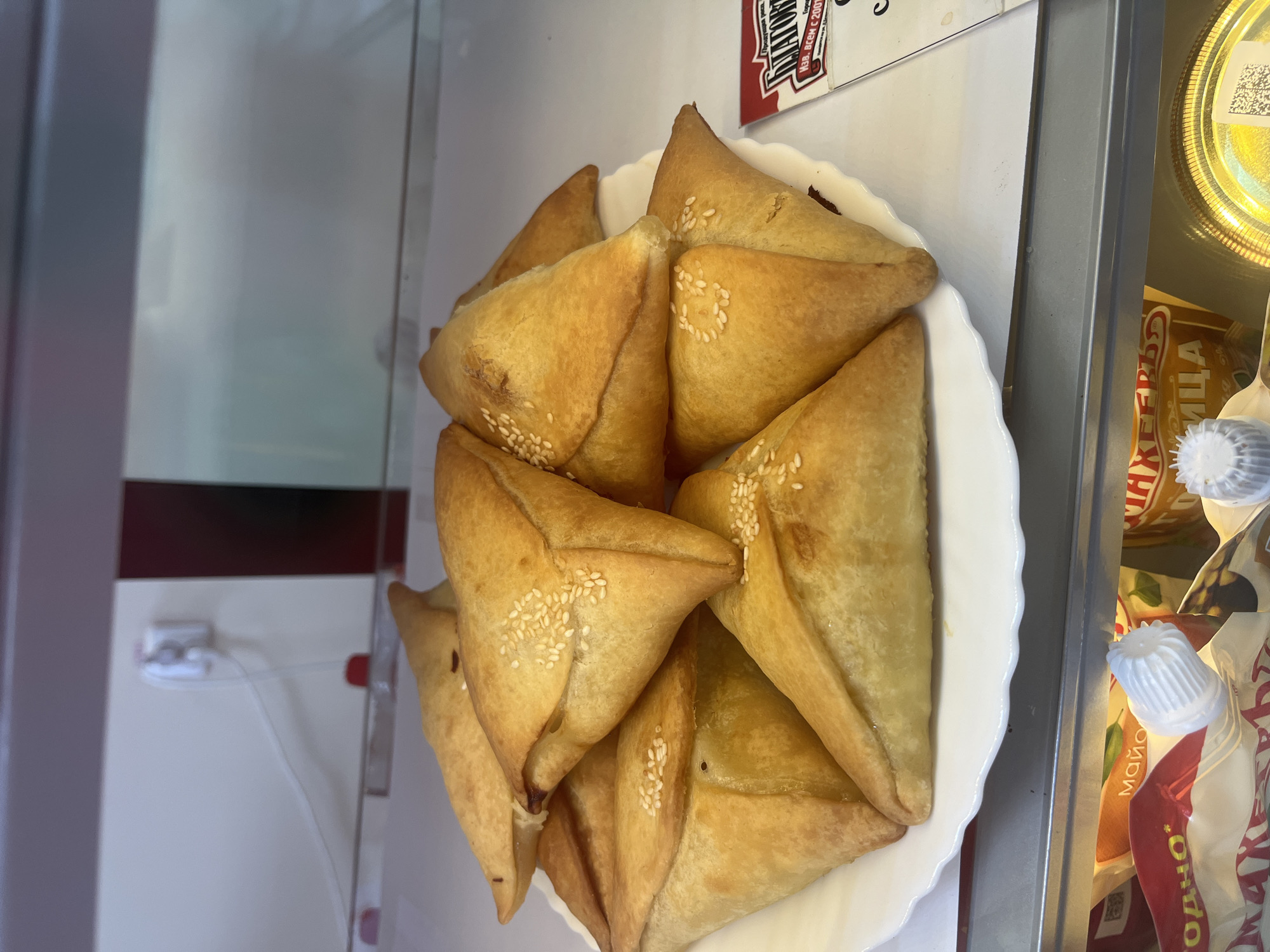 A white plate filled with golden-brown, triangular pastries topped with sesame seeds is displayed in a refrigerated case. The pastries appear to be baked and have a slightly flaky texture. In the background, a white electrical outlet with a cord is visible on a light-colored wall. Below the plate, a shelf in the refrigerated case contains various food products with visible labels, including one with the word "МАЙО" (mayonnaise). A red and white sign with Cyrillic text is also present to the right of the pastries. The overall scene suggests a retail or food service environment.