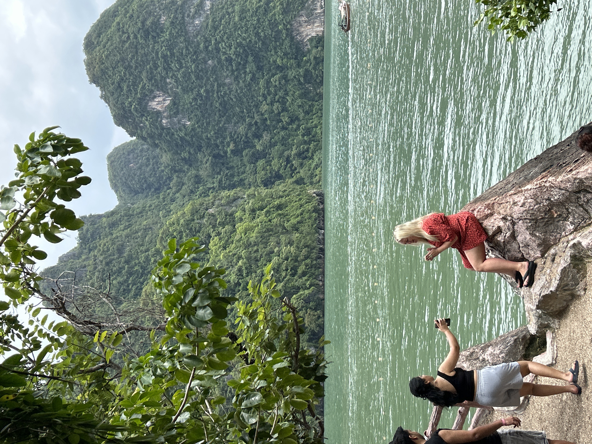 In Krasom, Thailand, two women and a man stand by the sea. One woman, wearing a black crop top and gray shorts, holds a drink and turns towards the other woman. The second woman, with blonde hair, is seated on a rock, wearing a red dress and holding a drink. The man, partially visible on the left, wears a black tank top and shorts. The background features lush green mountains and a boat on the turquoise water.