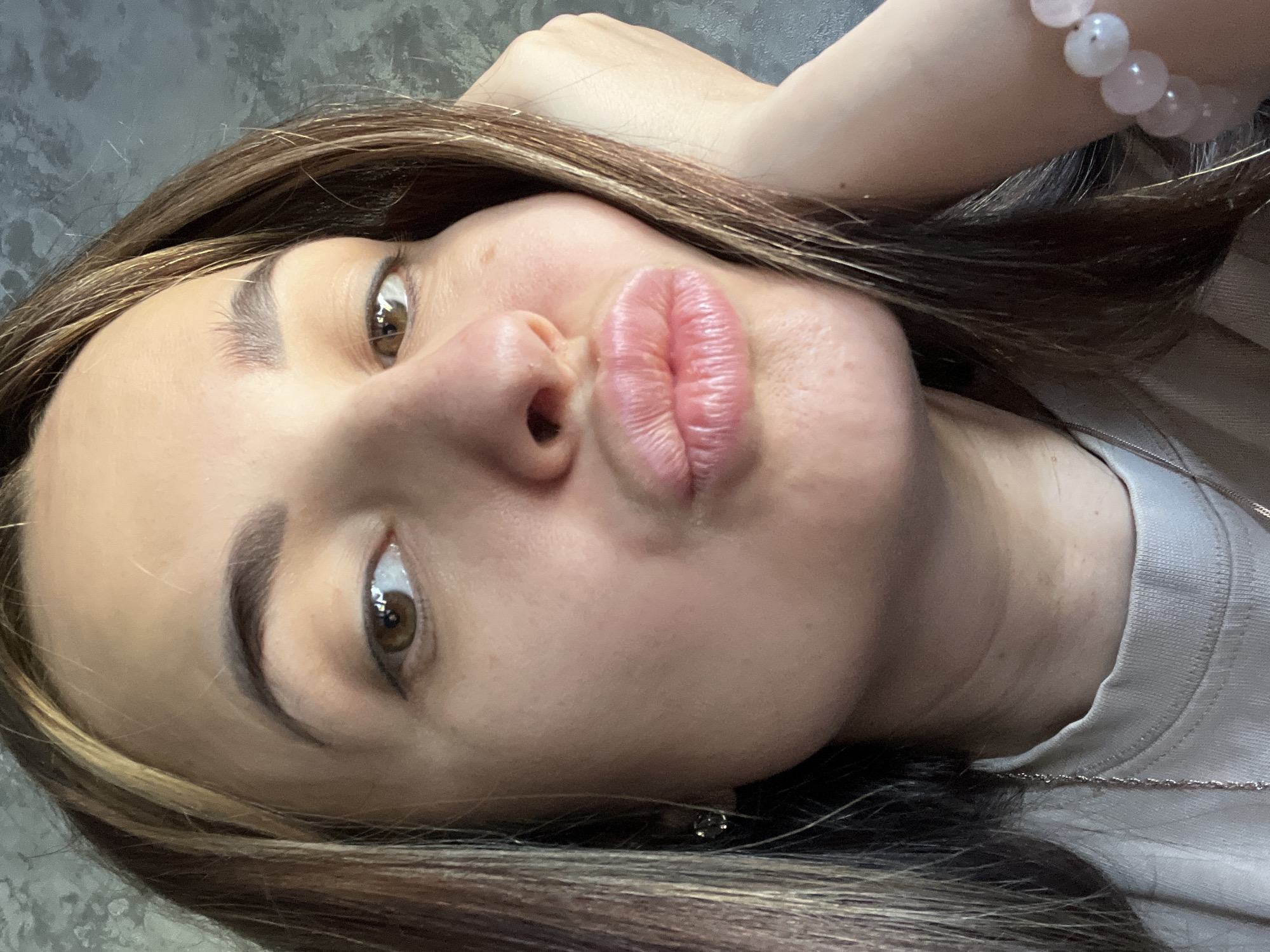 A close-up selfie depicts a young woman with light brown hair with blonde highlights, brown eyes, and full lips puckered in a kiss. She is wearing a light-colored shirt and a bracelet with pink beads on her wrist, which is resting near her head. The background is a textured grey wall. Location: Unknown, Unknown.