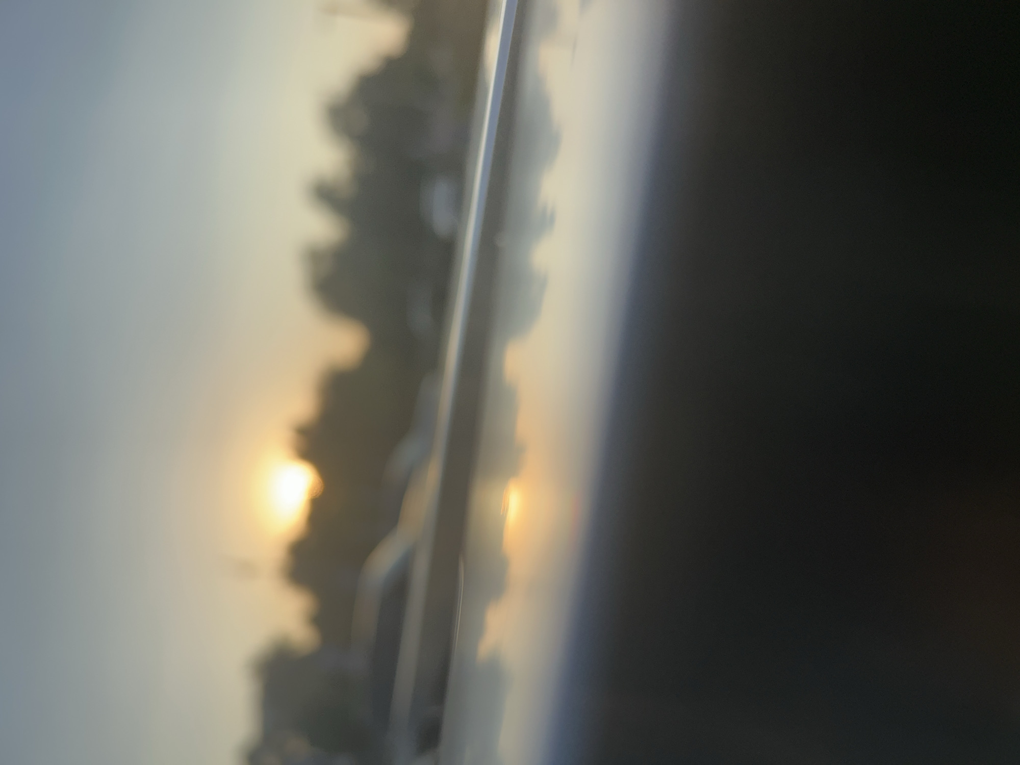 The image is a blurred, low-angle shot of a sunset or sunrise. The sun, a bright, hazy orb, is visible in the upper left portion of the frame. Silhouetted trees and possibly buildings form a dark line against the horizon. In the foreground, a reflective surface, possibly the hood of a car, mirrors the sky and the sun. The overall impression is one of a peaceful, natural scene, possibly taken from a vehicle. There are no people or specific landmarks visible that would allow for pinpointing a location within Krasnodar, Russia.
