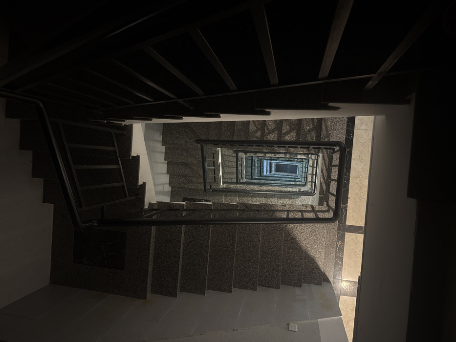 This image depicts a residential stairwell, viewed from an elevated position looking directly down into a multi-story, open central shaft, within Ngũ Hành Sơn Ward, Vietnam. The stairwell consists of square or U-shaped flights of stairs constructed from dark, speckled granite-like material, bordered by black metal railings with horizontal balusters. The scene is dimly lit, with the immediate foreground and sections of the upper right appearing almost entirely dark. Natural or artificial light illuminates the deeper sections of the stairwell, particularly within the central void, creating a repeating pattern of light and shadow on the descending steps and walls. The lowest visible point exhibits a rectangular area of brighter illumination, suggesting a light source or an exit. No individuals or personal effects are present.