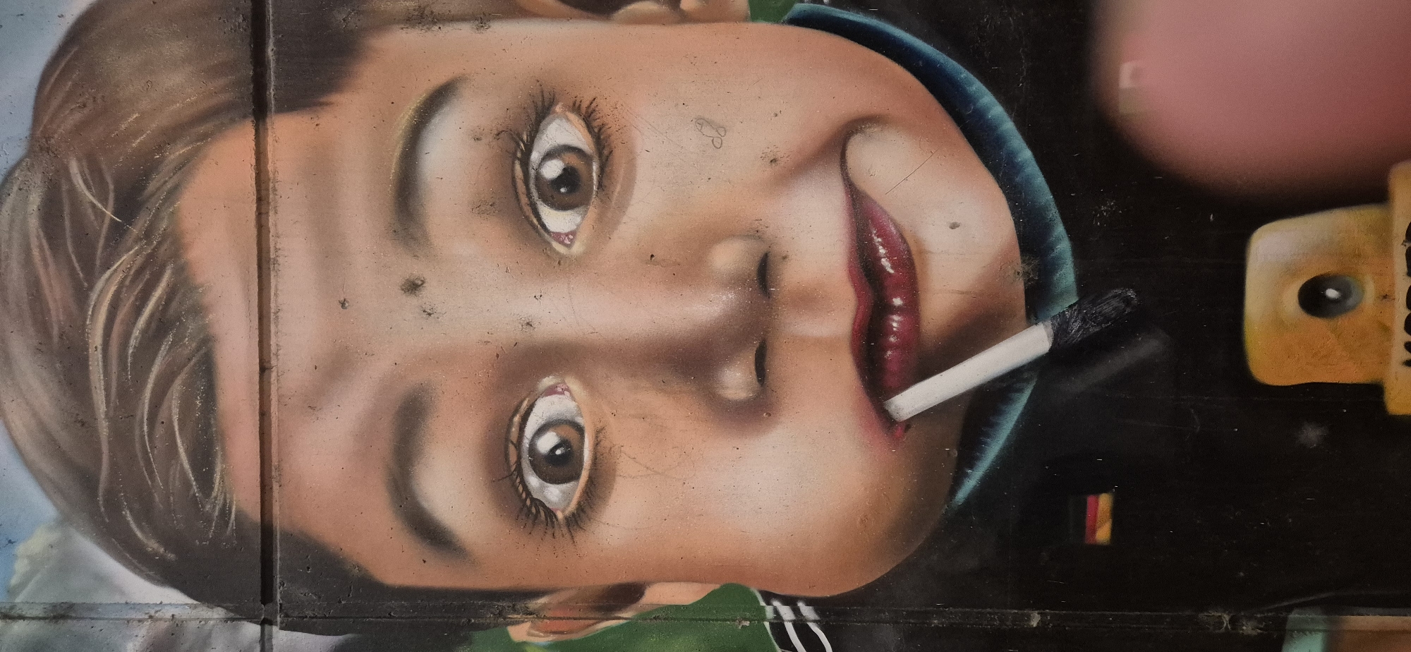 The image is a close-up, low-angle photograph of a graffiti mural depicting a child's face. The mural is on a concrete wall, with visible horizontal lines indicating the segmented construction of the wall. The child has puffed cheeks, wide eyes with dilated pupils, and dark eyebrows. A white cigarette is placed in the child's mouth. The child is wearing a dark garment with a teal collar, and a small German flag is visible on the left side of the garment. In the lower part of the image, part of a yellow spray paint can nozzle and a blurred pink object, possibly a finger, are visible. The graffiti style is realistic with shading and detail. The context of Budenheim, Germany is not directly visible in the image itself.