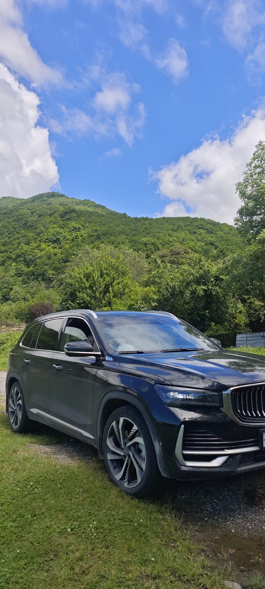 A black SUV is parked on a dirt path next to a grassy area, set against a picturesque mountainous landscape. The scene, located in Bzypta, Abkhazia, features lush green hills and mountains covered in dense forest, stretching towards a bright blue sky dotted with fluffy white clouds.
The vehicle is a modern crossover with a prominent dark grille, sleek headlights, and distinctive silver and black alloy wheels. Silver trim is visible around the windows and along the lower body. The driver's side front window is partially open, revealing a person's head and shoulder faintly in the driver's seat. The foreground shows vibrant green grass with small white flowers, contrasting with the gravelly path.
The time of day appears to be sunny daytime, likely late morning or early afternoon, given the clear sky and bright illumination. The weather is fair and pleasant. The overall setting suggests a tranquil, natural environment, possibly a stop during a road trip or an outdoor excursion. No specific activity other than the car being parked is depicted, and no visible text is present.