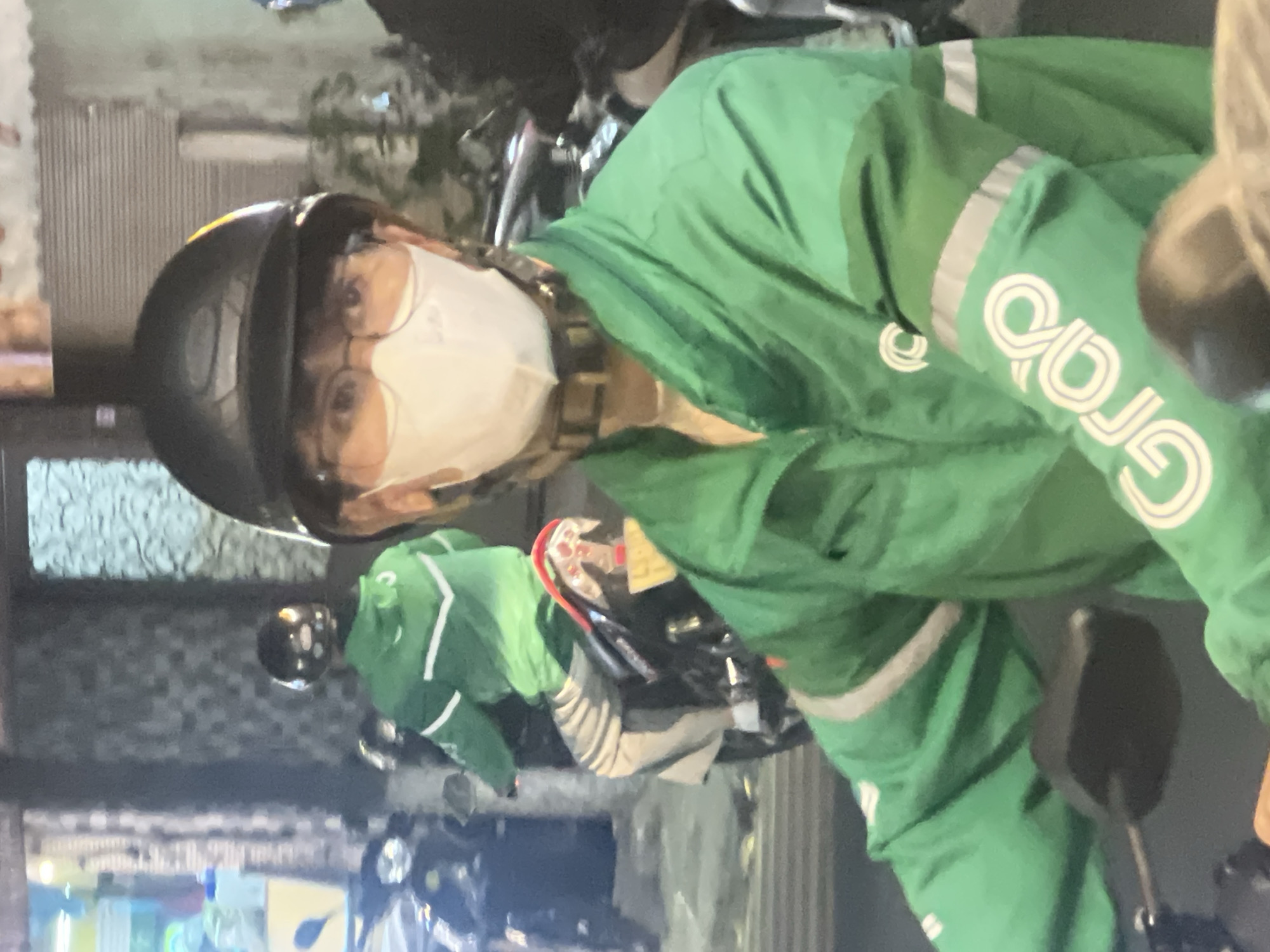 The image shows a person wearing a green jacket and a black helmet, riding a motorcycle in Đà Nẵng, Vietnam. The person is wearing a white face mask and glasses. The green jacket has white stripes on the sleeves and a logo on the chest. The motorcycle is black. The background is blurred, but it appears to be a street scene with buildings and other vehicles. The lighting suggests it is nighttime.