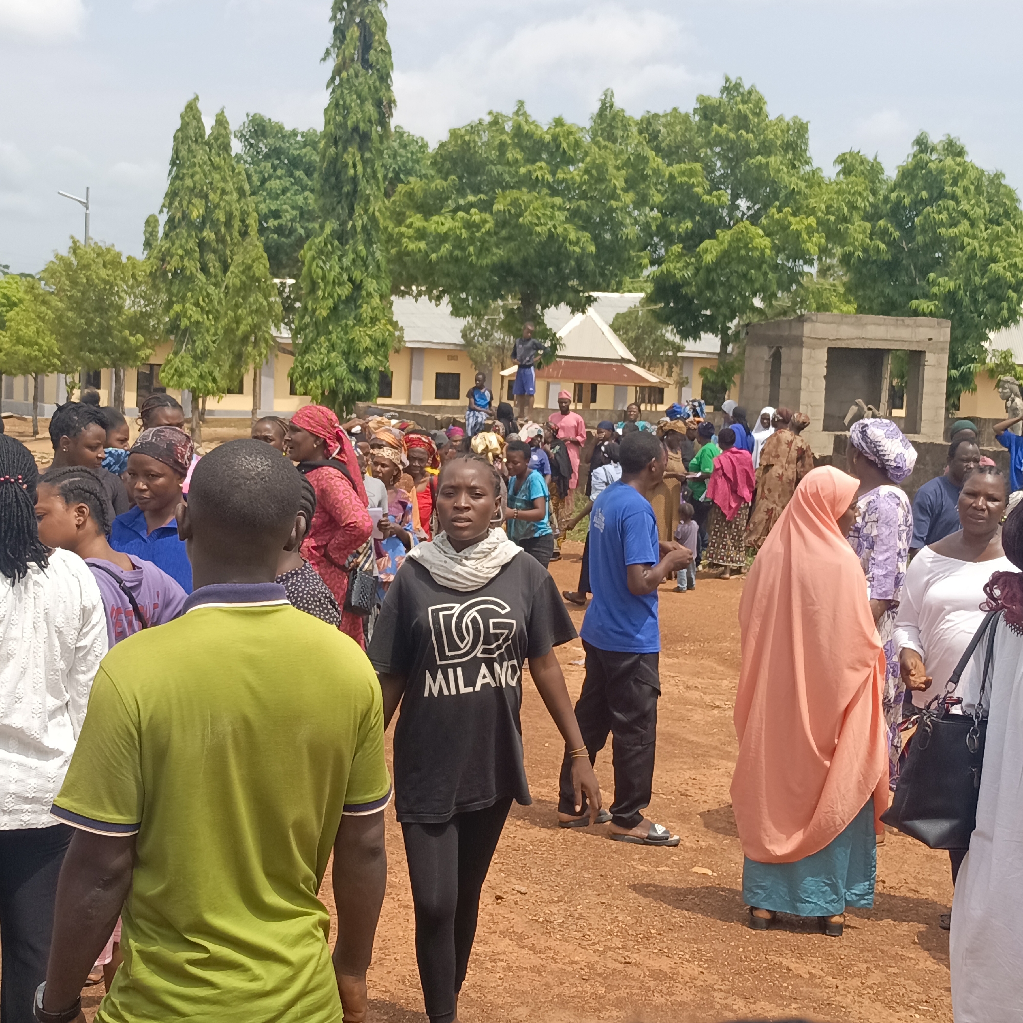In an outdoor setting in Akwanga, Nigeria, a diverse group of people, predominantly women, are gathered in a sandy area with buildings and tall trees in the background. Individuals are engaged in various activities, including walking, standing, and interacting with each other. The attire ranges from casual t-shirts and trousers to traditional garments, including headscarves and hijabs. Incomplete concrete structure visible in the background.